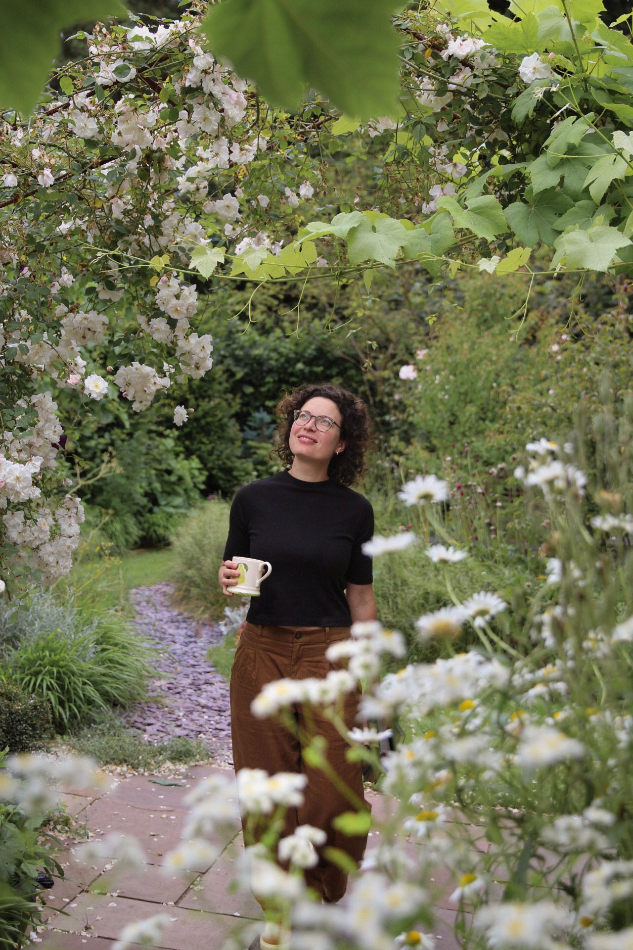A woman holding a mug walks through a lush garden archway covered in white climbing roses.
