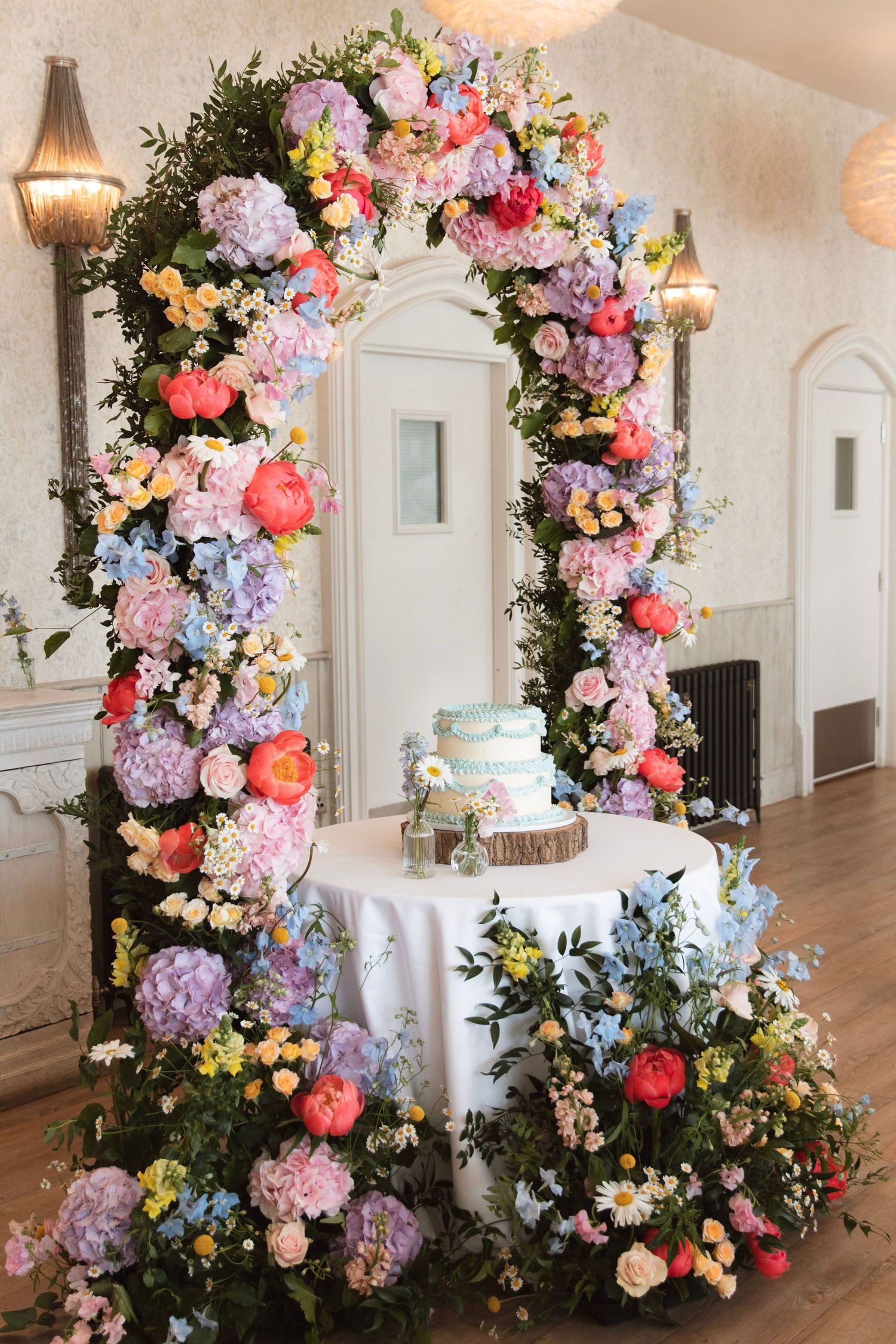 Colorful floral arch with pink, blue, and coral flowers framing a wedding cake table in an elegant white venue.
