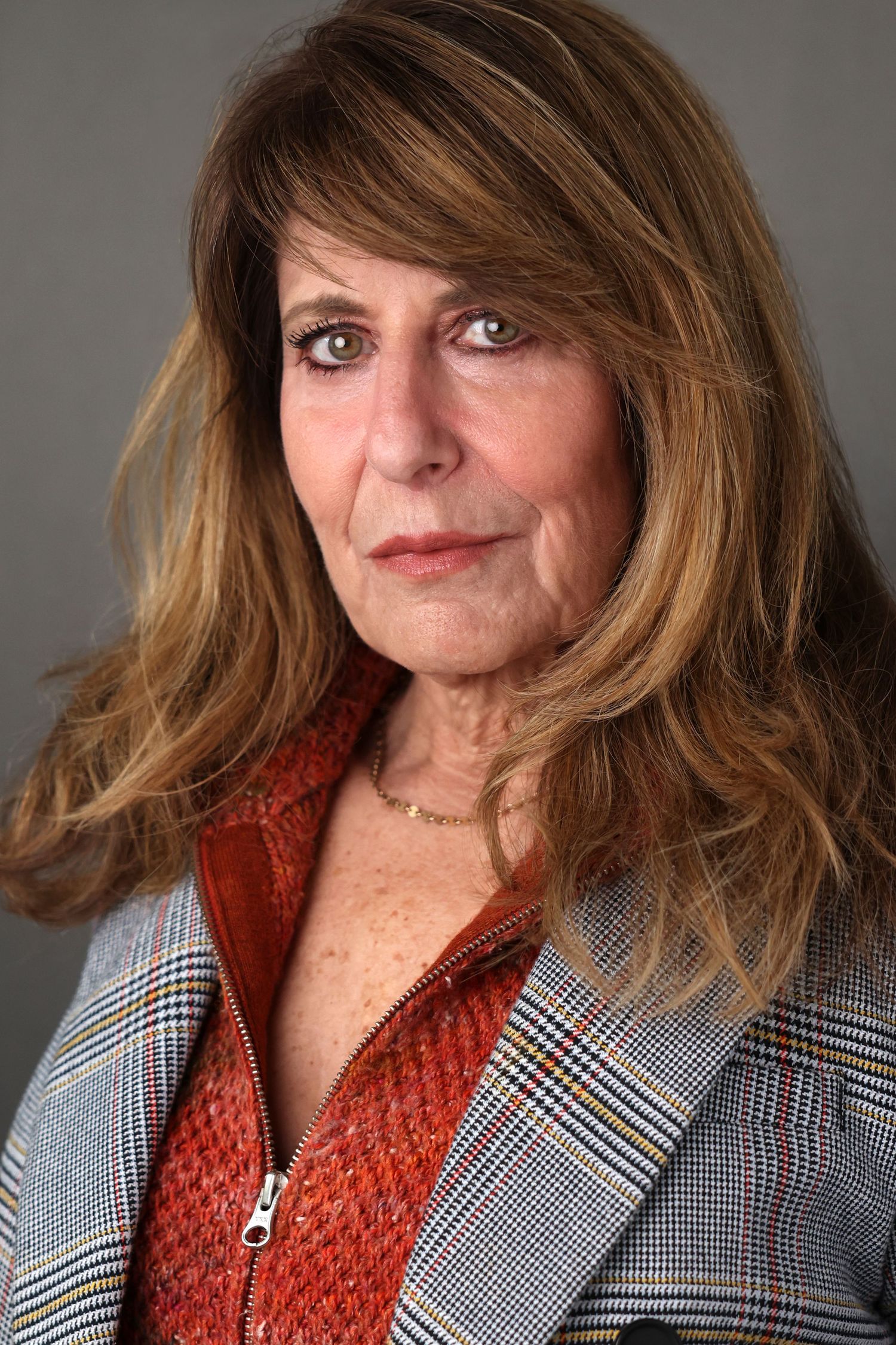 Portrait of a woman with long wavy auburn hair wearing a rust zip-up top and gray plaid blazer against a neutral gray background.