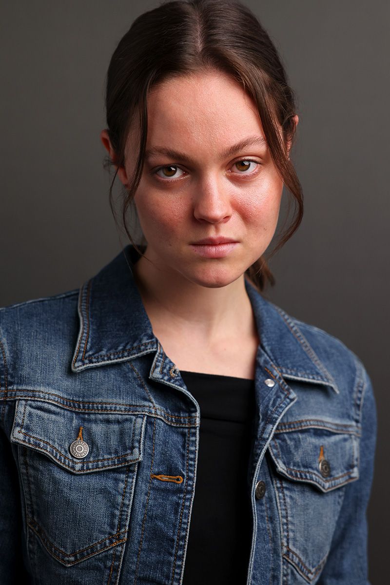 Young woman with freckles in a denim jacket poses against a gray background with a serious expression.