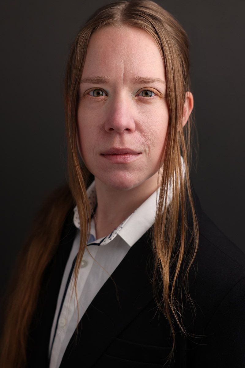 Professional headshot of a woman with long straight hair wearing a black blazer and white shirt against dark background.