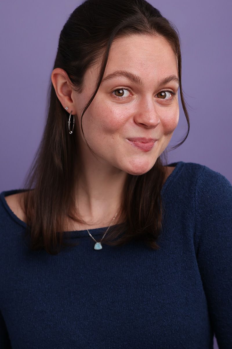 Young woman with dark hair wearing a blue sweater and teal pendant necklace, smiling playfully against purple background.