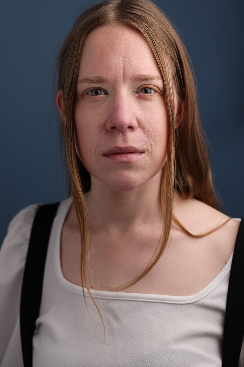 A young woman with long blonde hair wearing a white top and suspenders, looking sad against a blue-gray background.