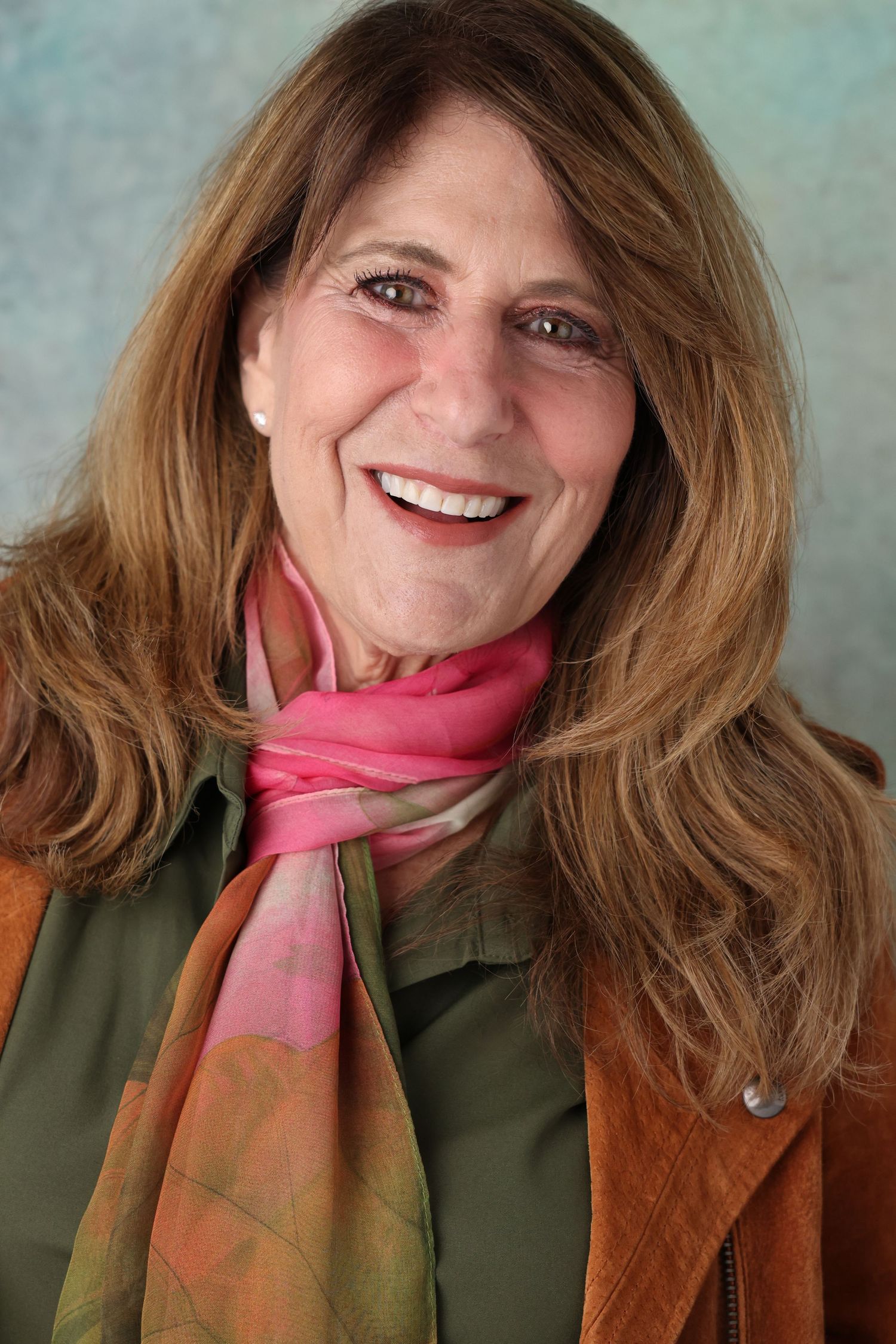 Smiling woman with long auburn hair wearing a pink and orange silk scarf, olive green top and rust suede jacket against a soft teal backdrop