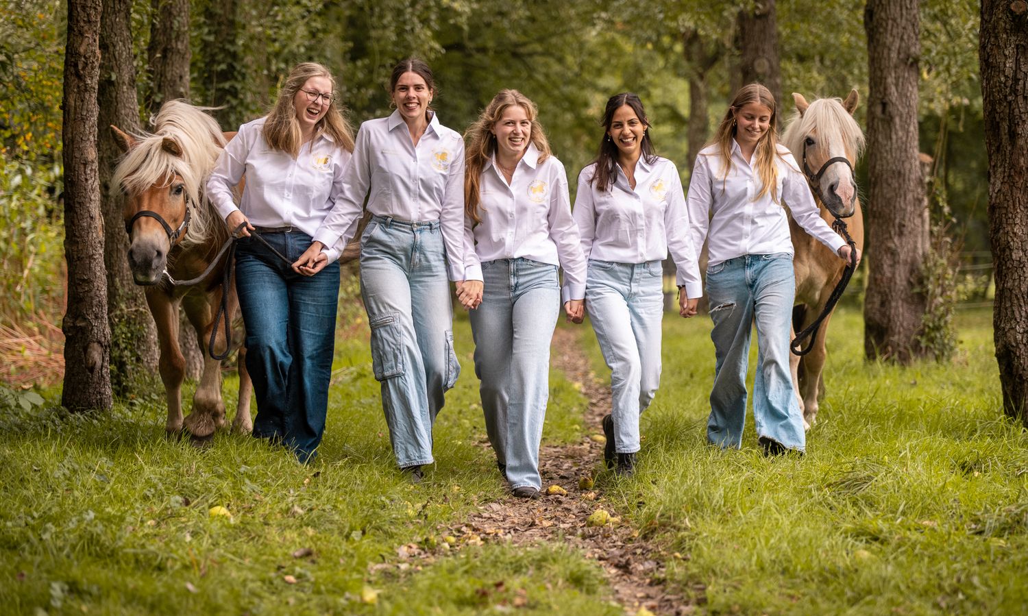 Five women in white shirts and jeans walk through a forest path with two horses in a lush green setting.