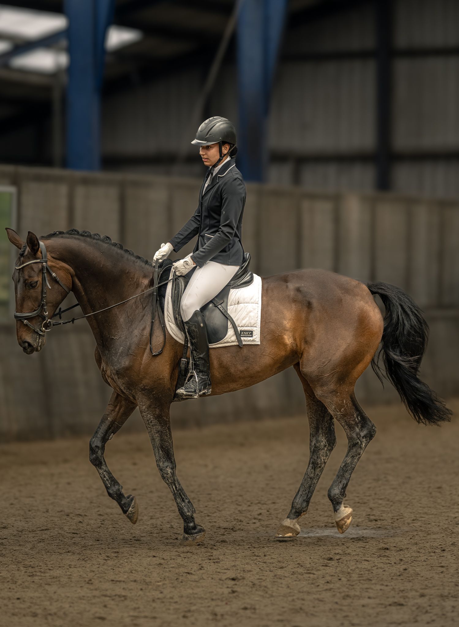 A rider in a black jacket and helmet performs dressage on a bay horse in an indoor equestrian arena.