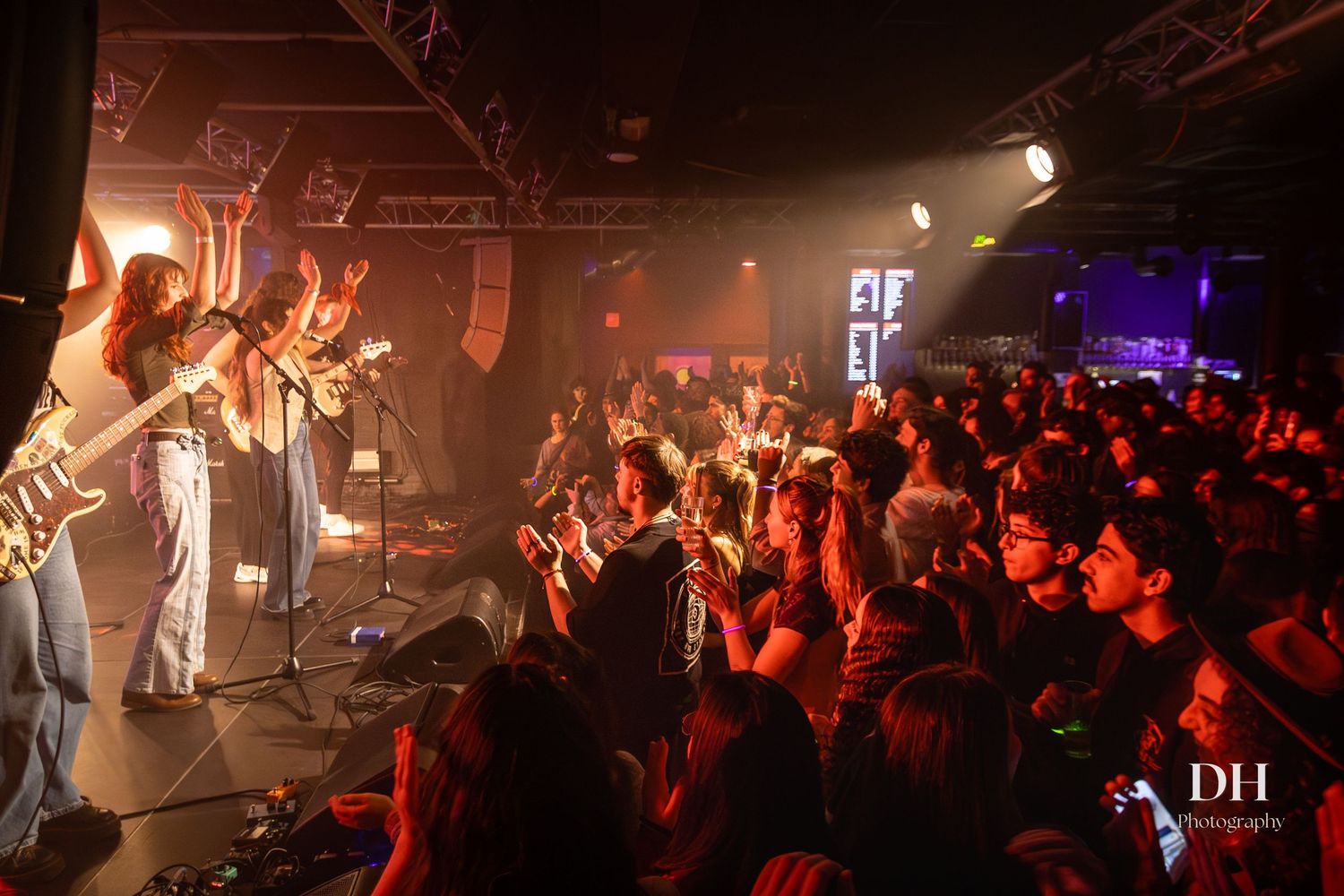 Band performing on stage to large enthusiastic crowd at energetic indoor live music concert.