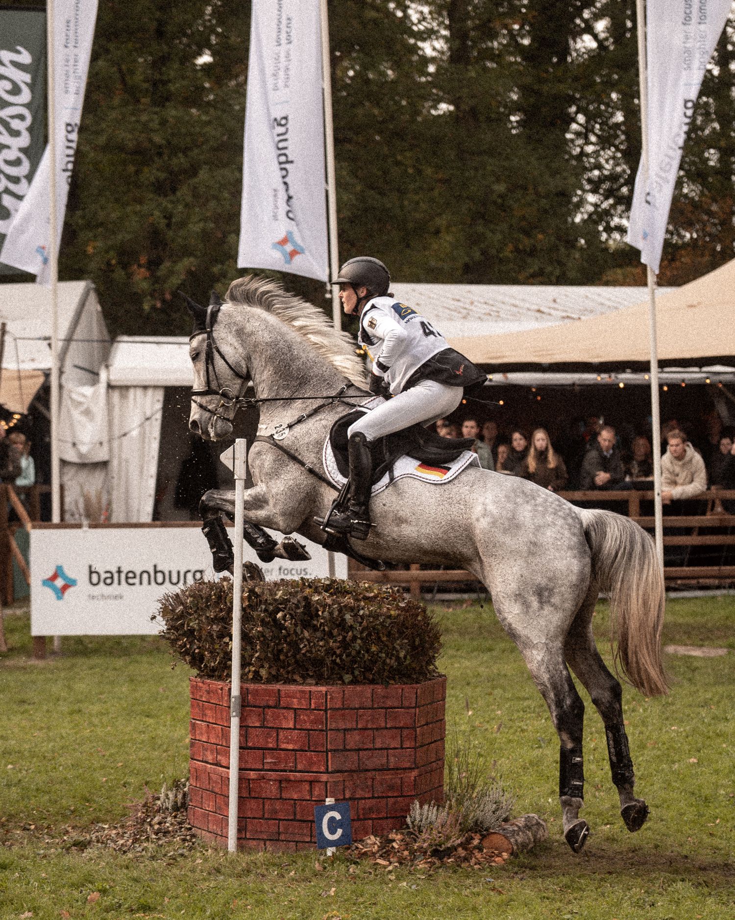 An equestrian on a grey horse jumps over a brick obstacle at an outdoor show jumping competition.