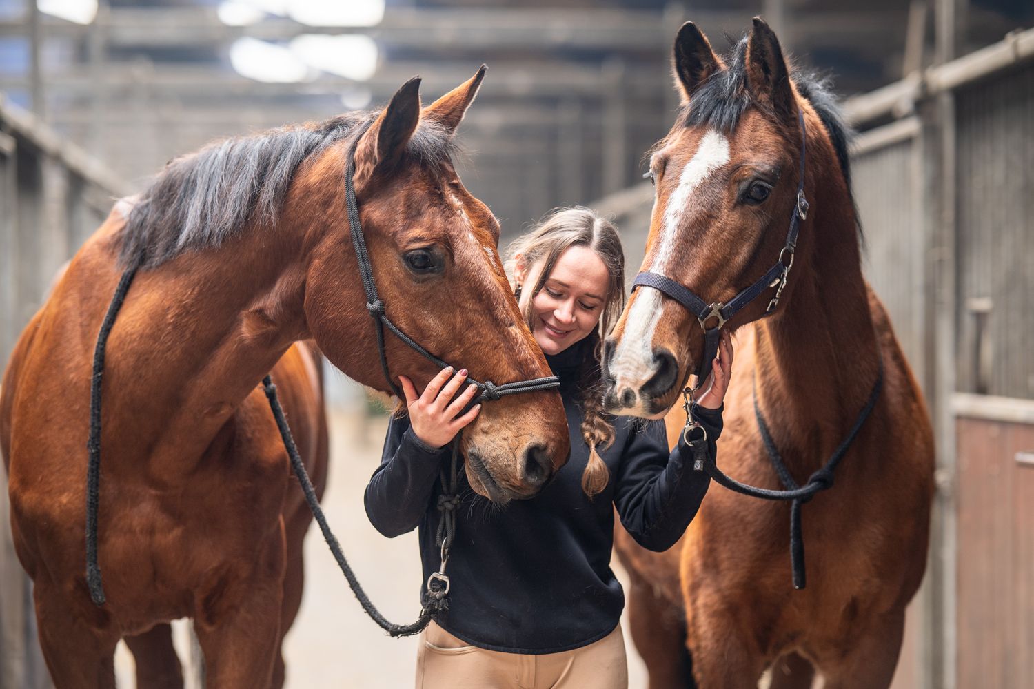 A young woman smiles warmly while hugging two chestnut horses inside a stable.