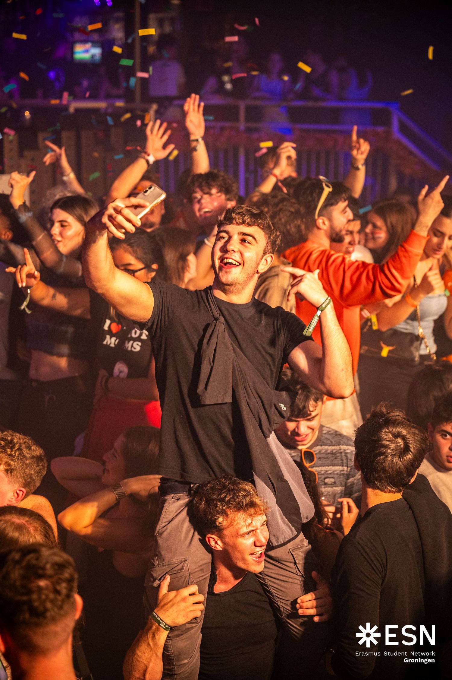 Smiling young man taking selfie on shoulders in energetic ESN party crowd with confetti falling.
