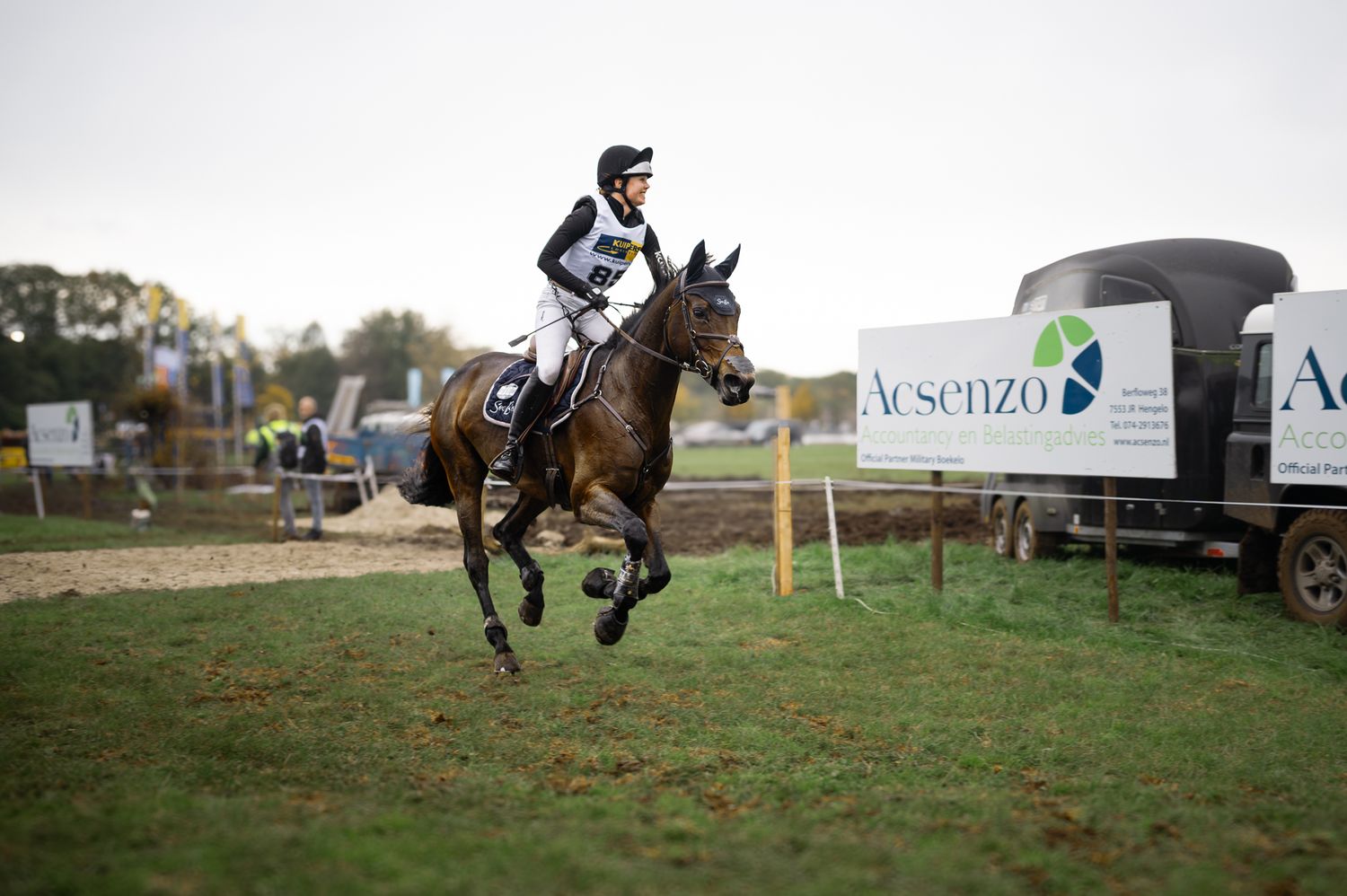 A horse and rider gallop past sponsor banners during an outdoor equestrian cross-country event.