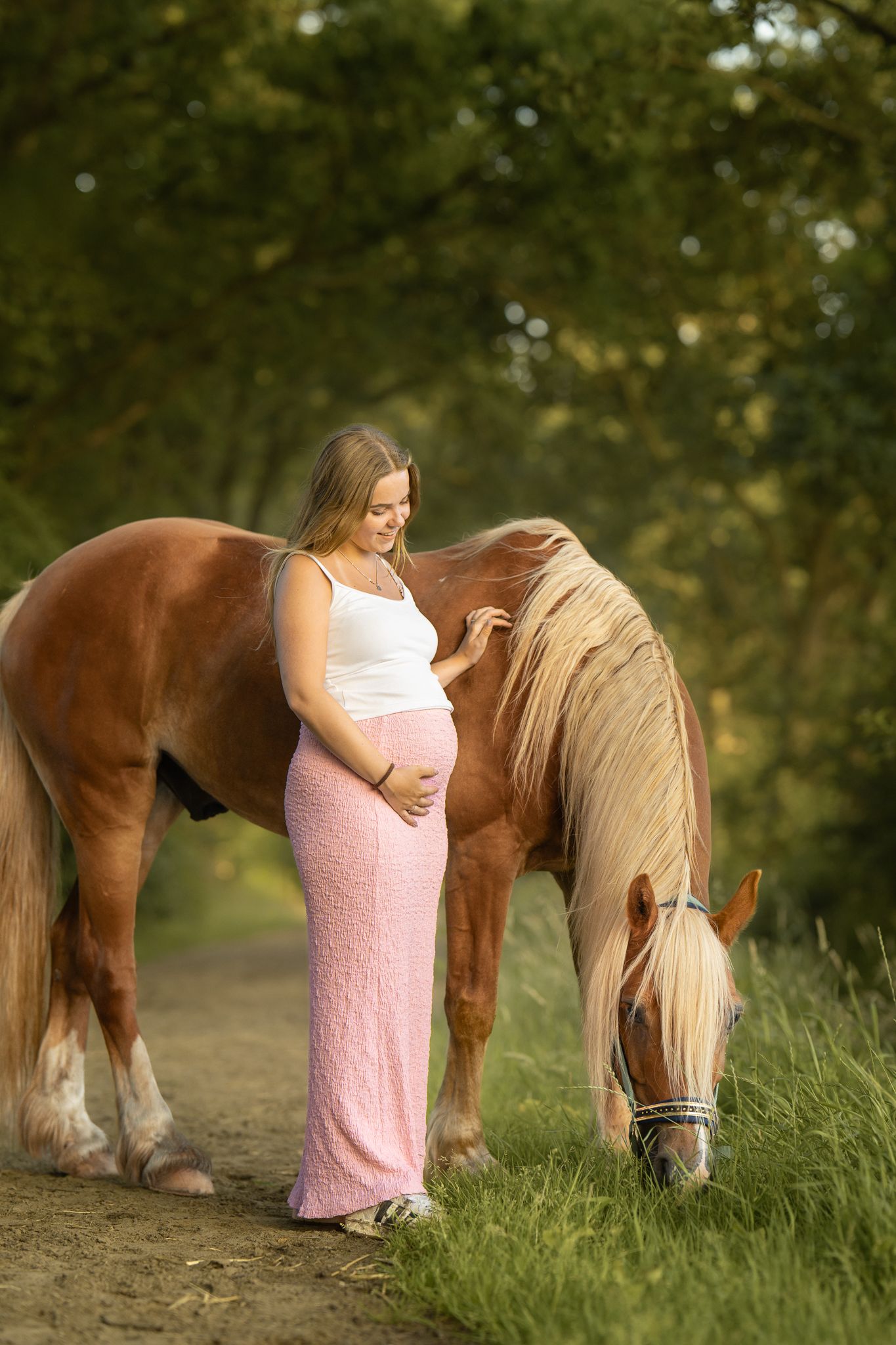 Pregnant woman in pink skirt standing beside a grazing chestnut horse outdoors.