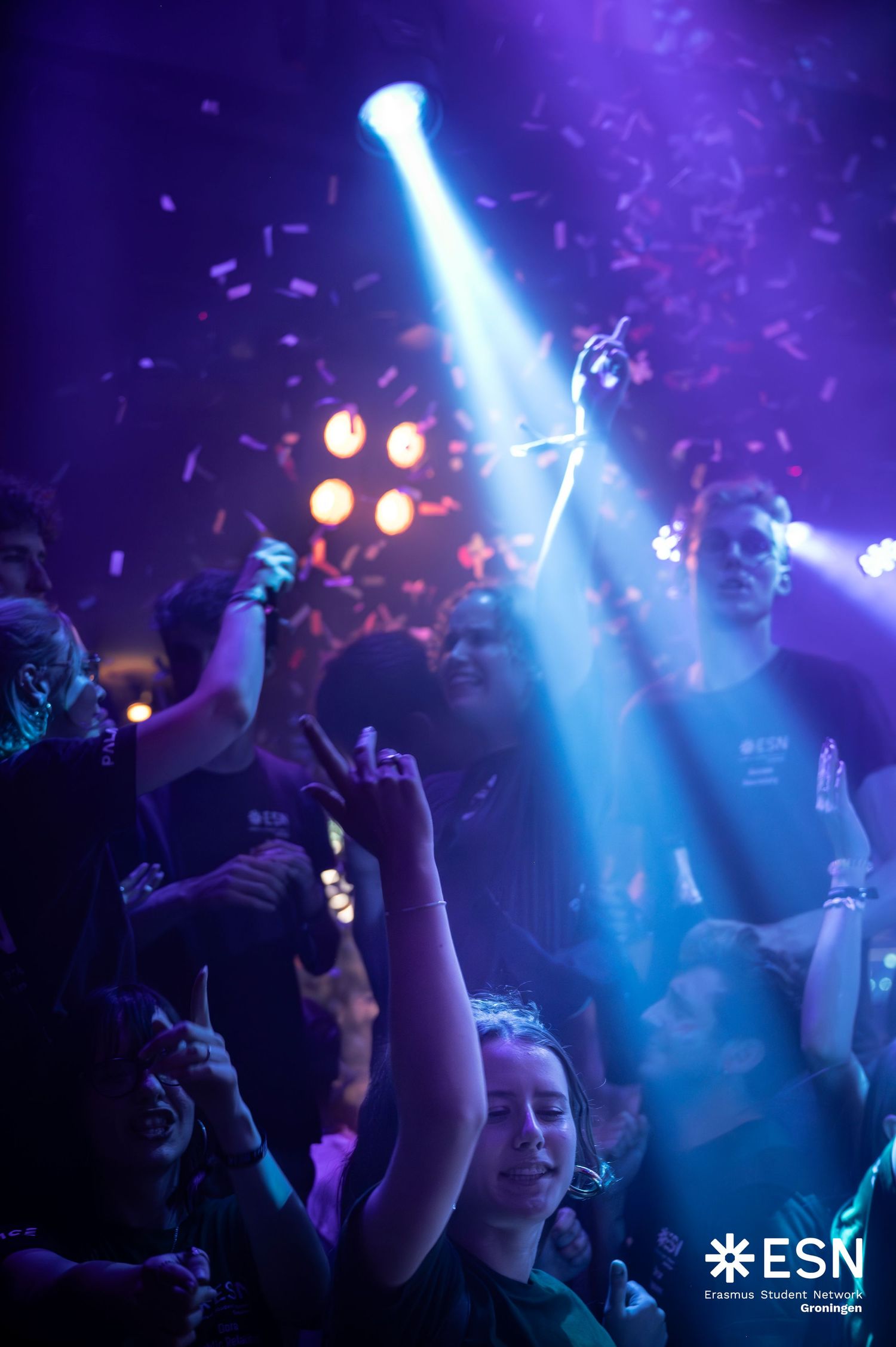 ESN party crowd with hands raised under bright blue spotlight and falling confetti in dark venue.