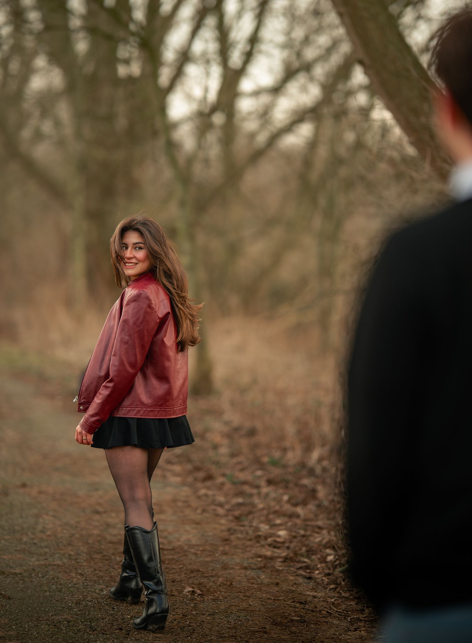 Young woman in red leather jacket smiling on forest path with person in foreground.