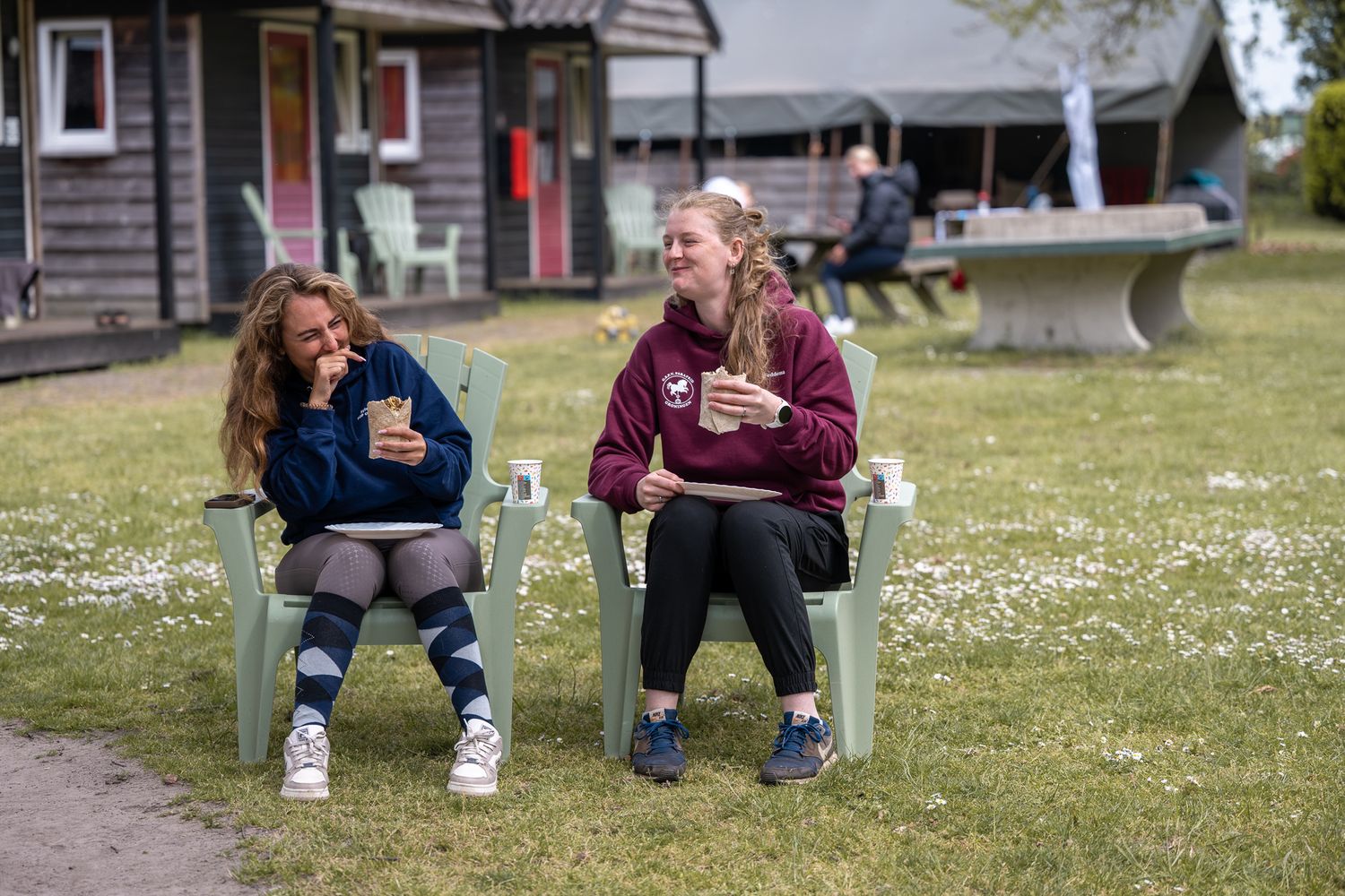 Two young women sit in green chairs outdoors, laughing and holding drinks on a sunny day.