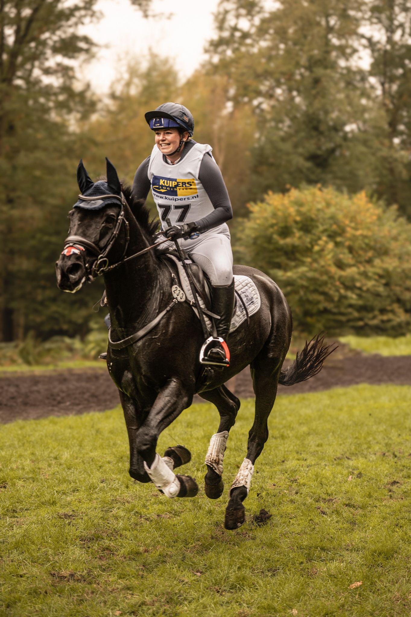 A smiling rider wearing number 77 gallops a black horse across a grassy cross-country course.