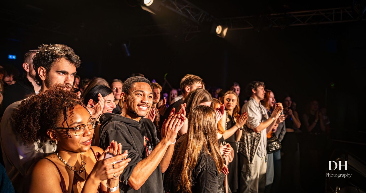 Diverse audience clapping and smiling at live music event under bright stage lights.
