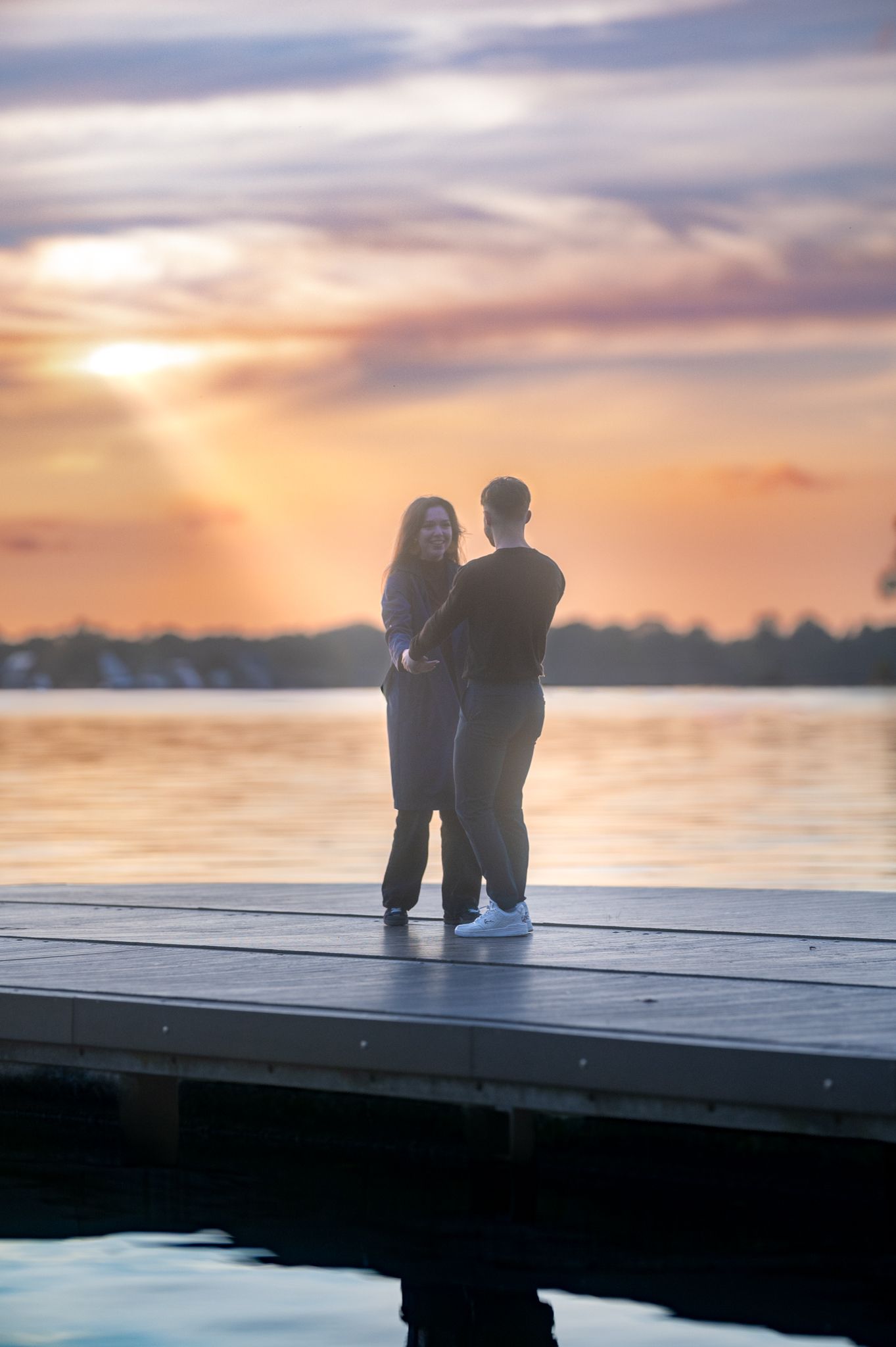 Couple embracing on a wooden dock at sunset over a calm lake with vibrant orange and purple skies.