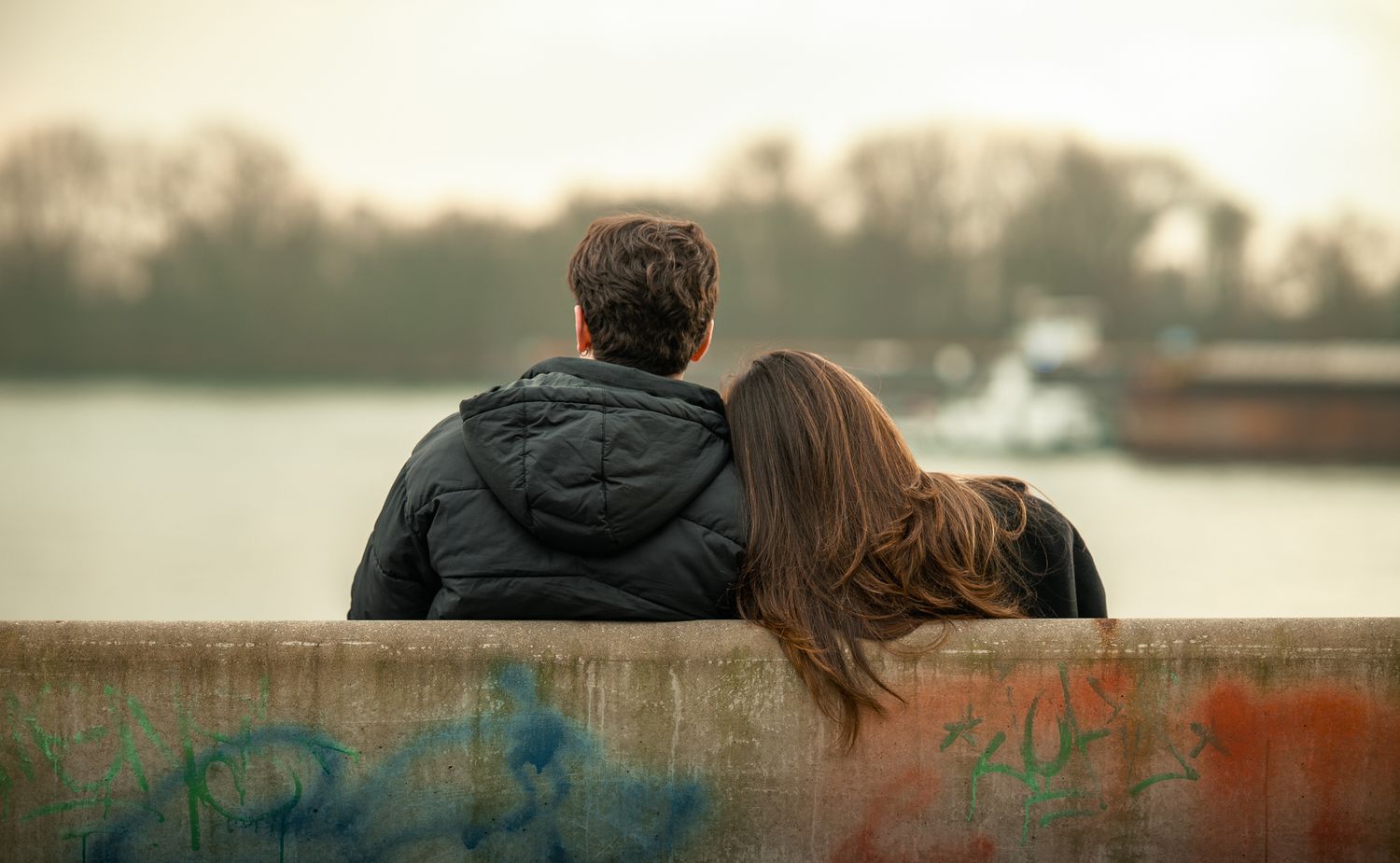 Romantic couple sitting together on a graffiti wall, leaning on each other while overlooking a calm river.