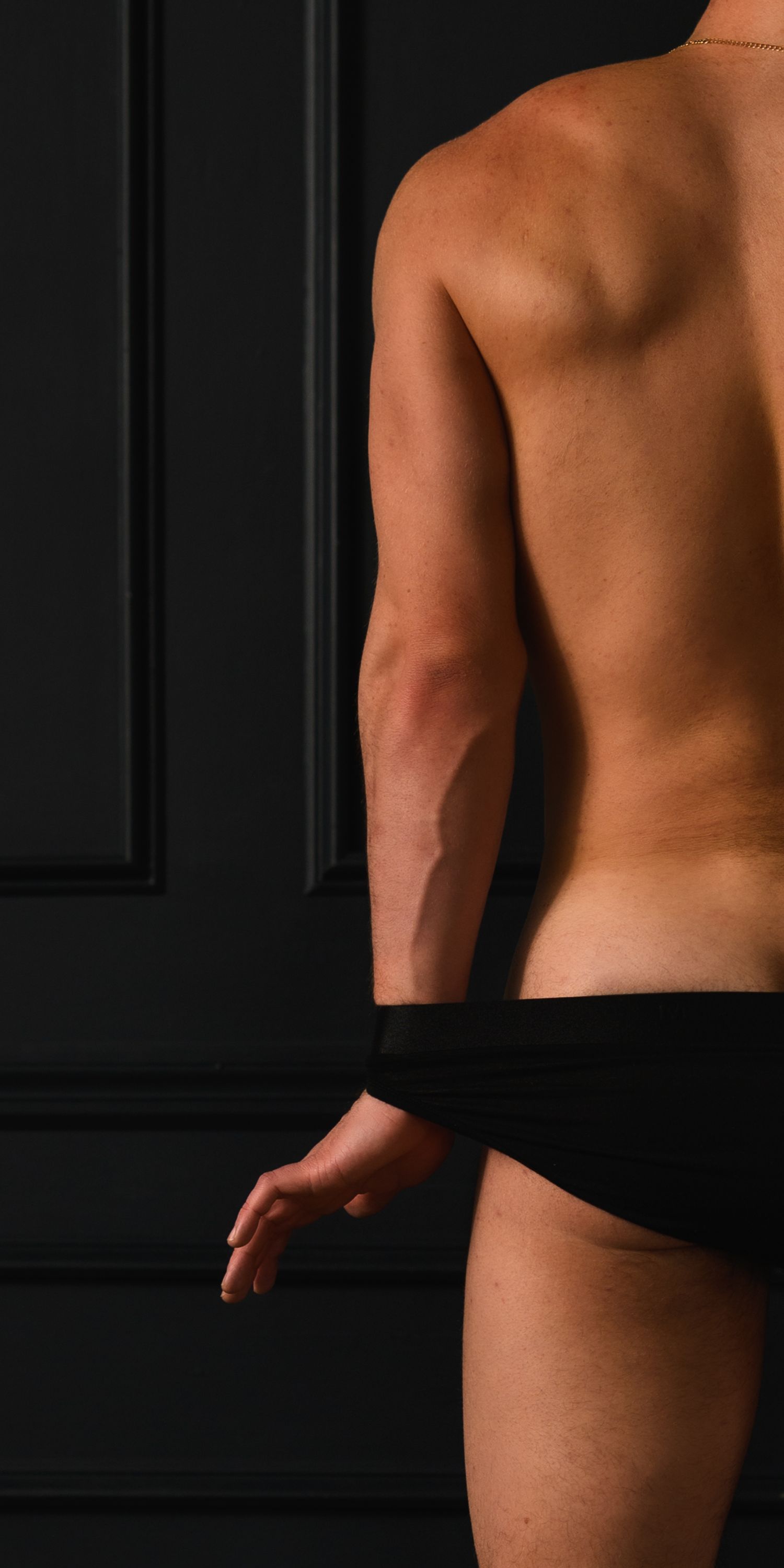 Side back view of a toned man in black briefs with a gold chain against a dark wall.