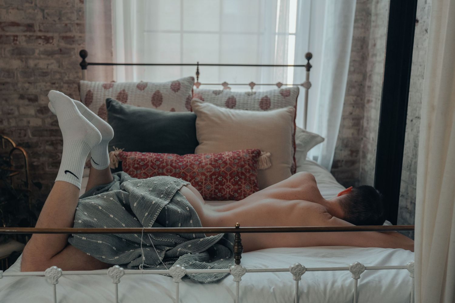 Person lying face down on a bed with colorful pillows near a window with white curtains, with a blanket covering his bare backside.