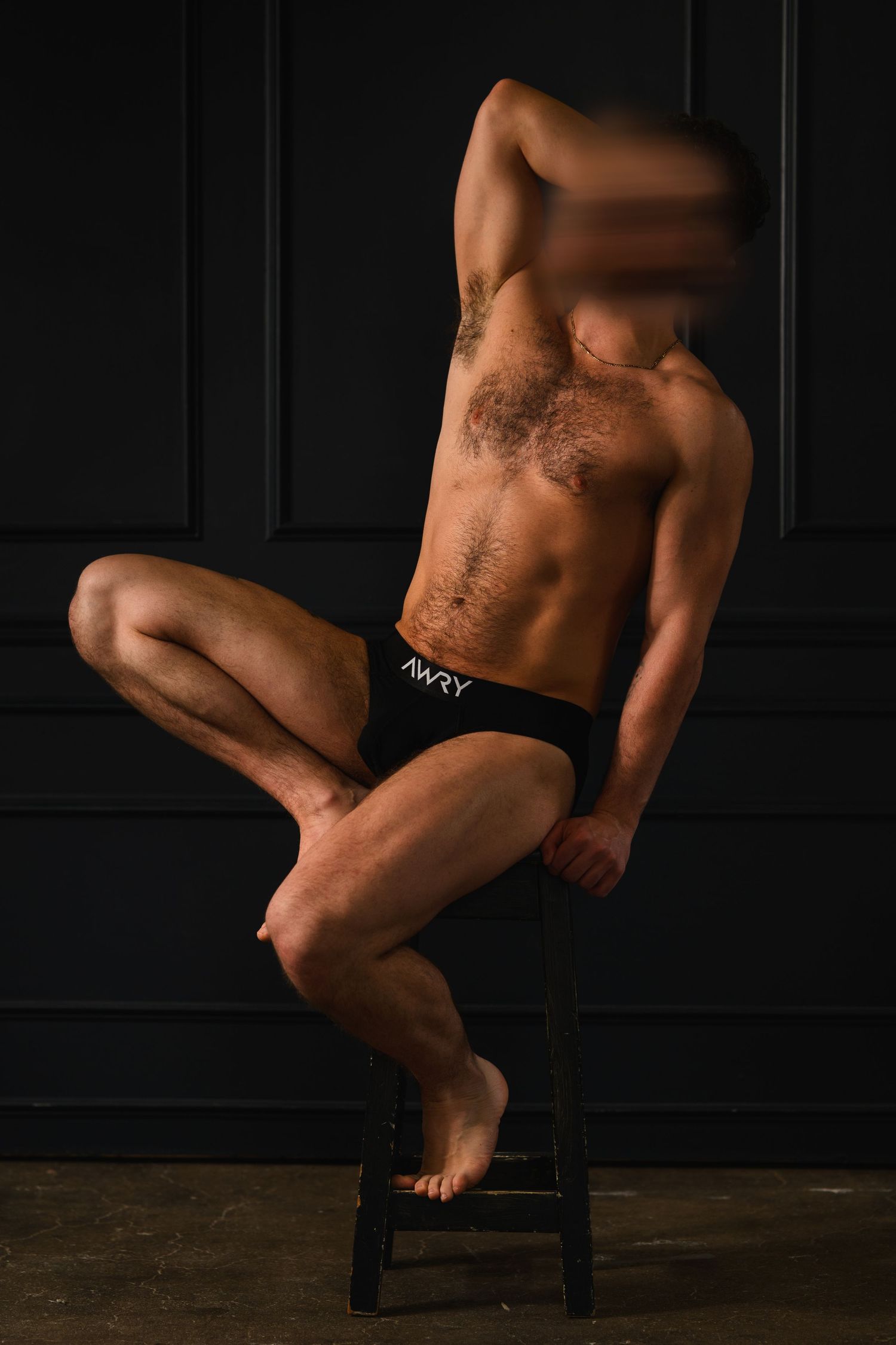 Muscular man posing on a stool wearing black AWRY briefs against a dark background.