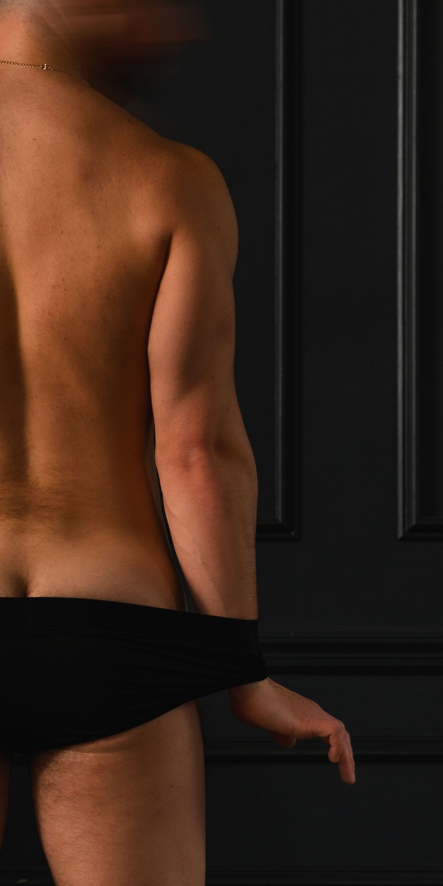 Side back view of a toned man in black briefs with a gold chain against a dark wall.