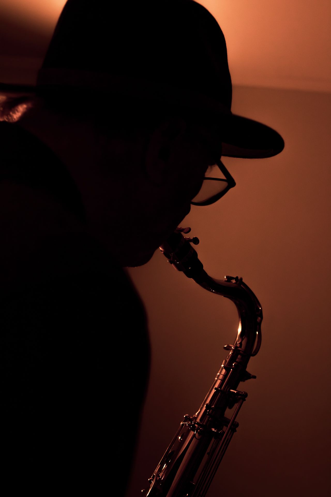 Silhouette of a jazz musician wearing a cap and glasses playing saxophone against a warm orange background.