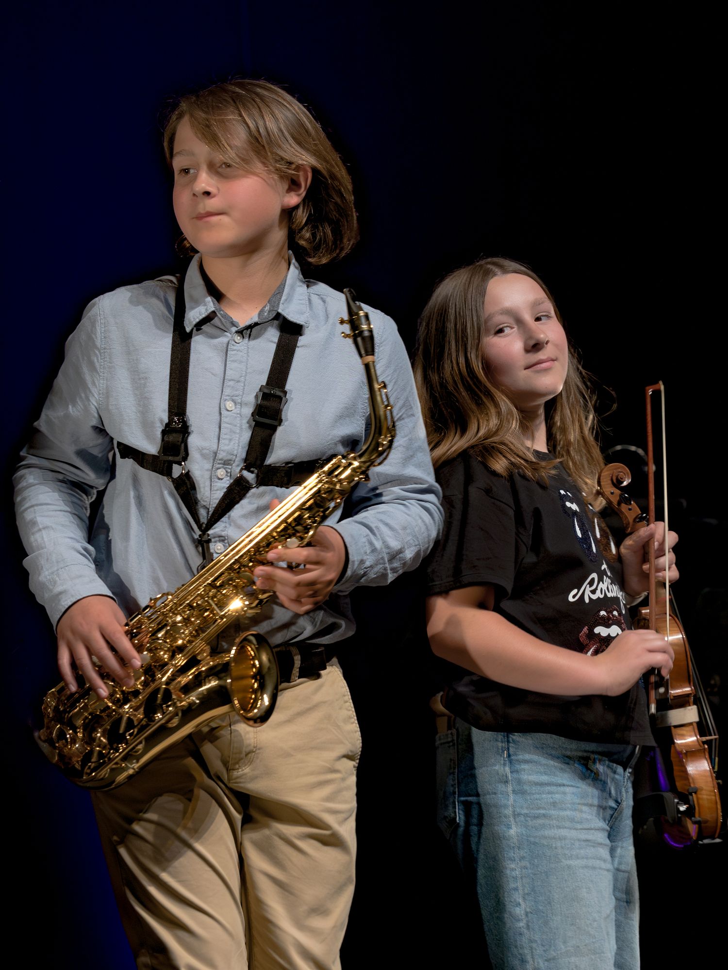 Two young musicians  boy playing saxophone and girl holding violin under stage lights.