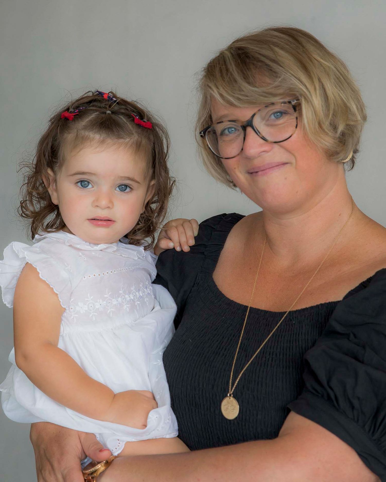 Woman with blonde bob and glasses holding young curly-haired toddler girl in white dress.
