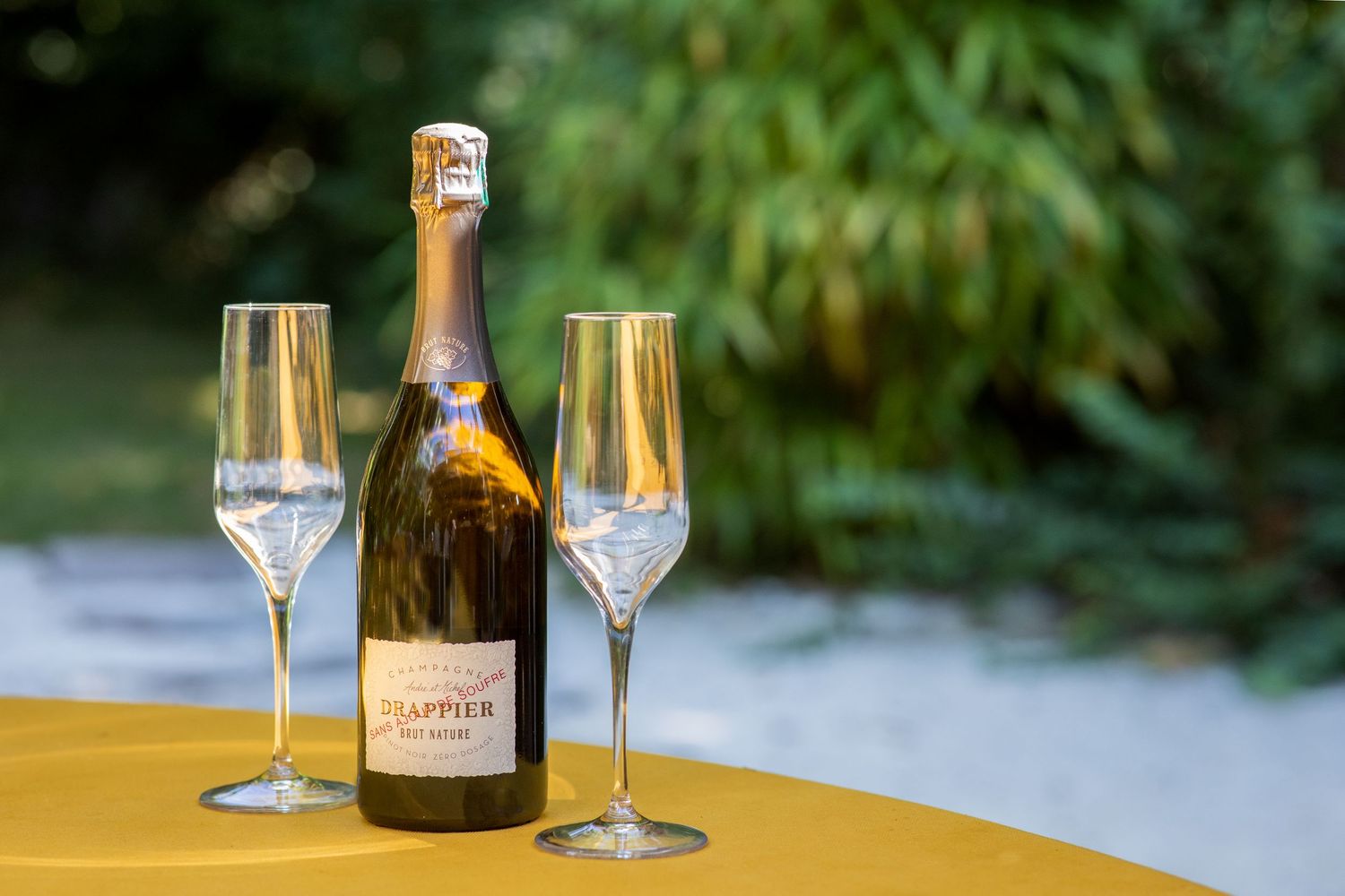 A bottle of Drappier champagne with two empty flutes on a yellow tablecloth with green foliage background.