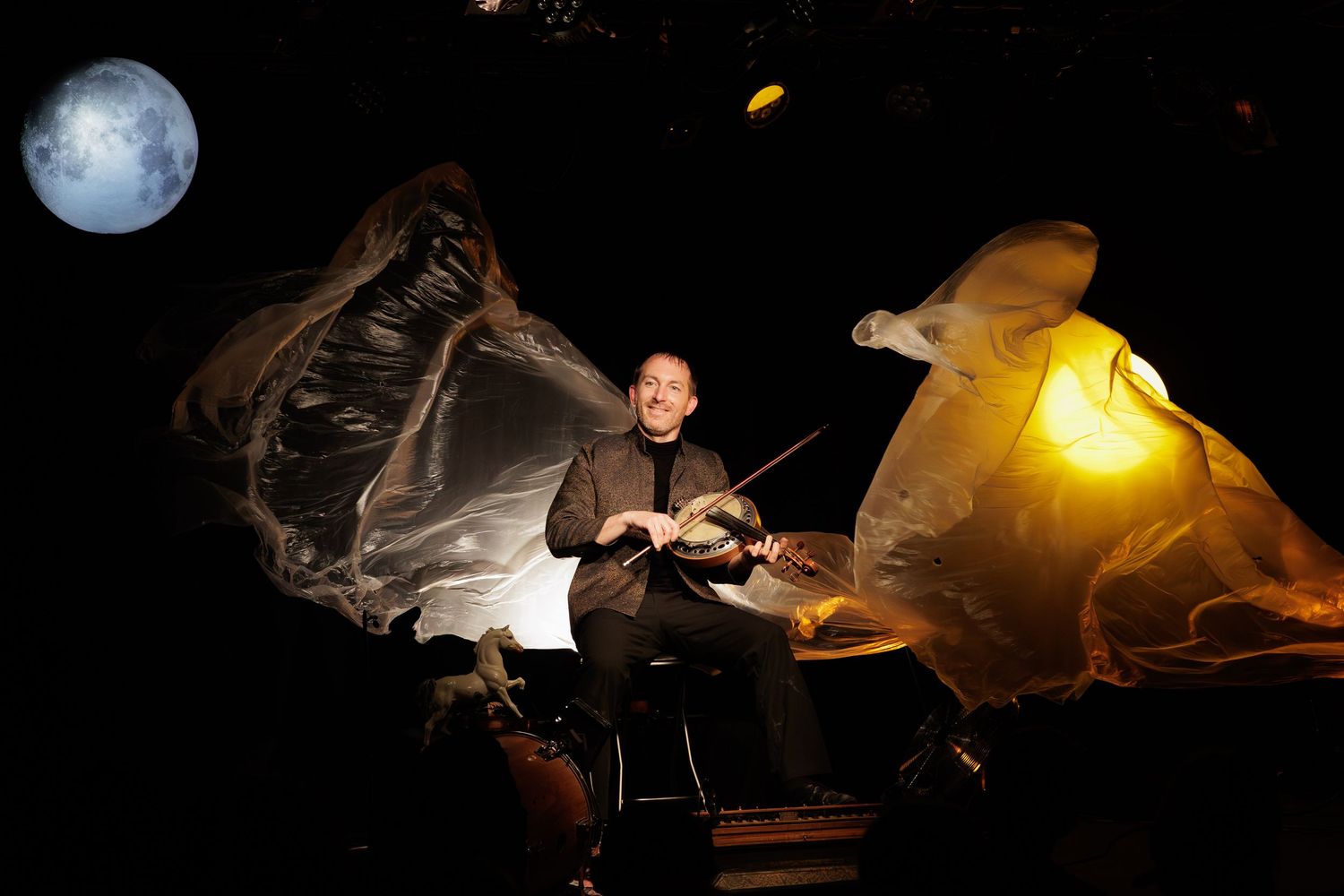 Dramatic theatrical performance with violinist playing amid flowing fabric and moonlit stage lighting.