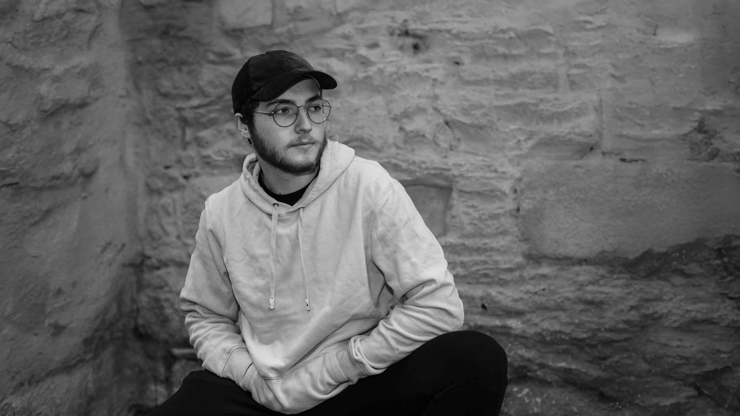 Black and white portrait of a young man in cap and glasses sitting against a stone wall.