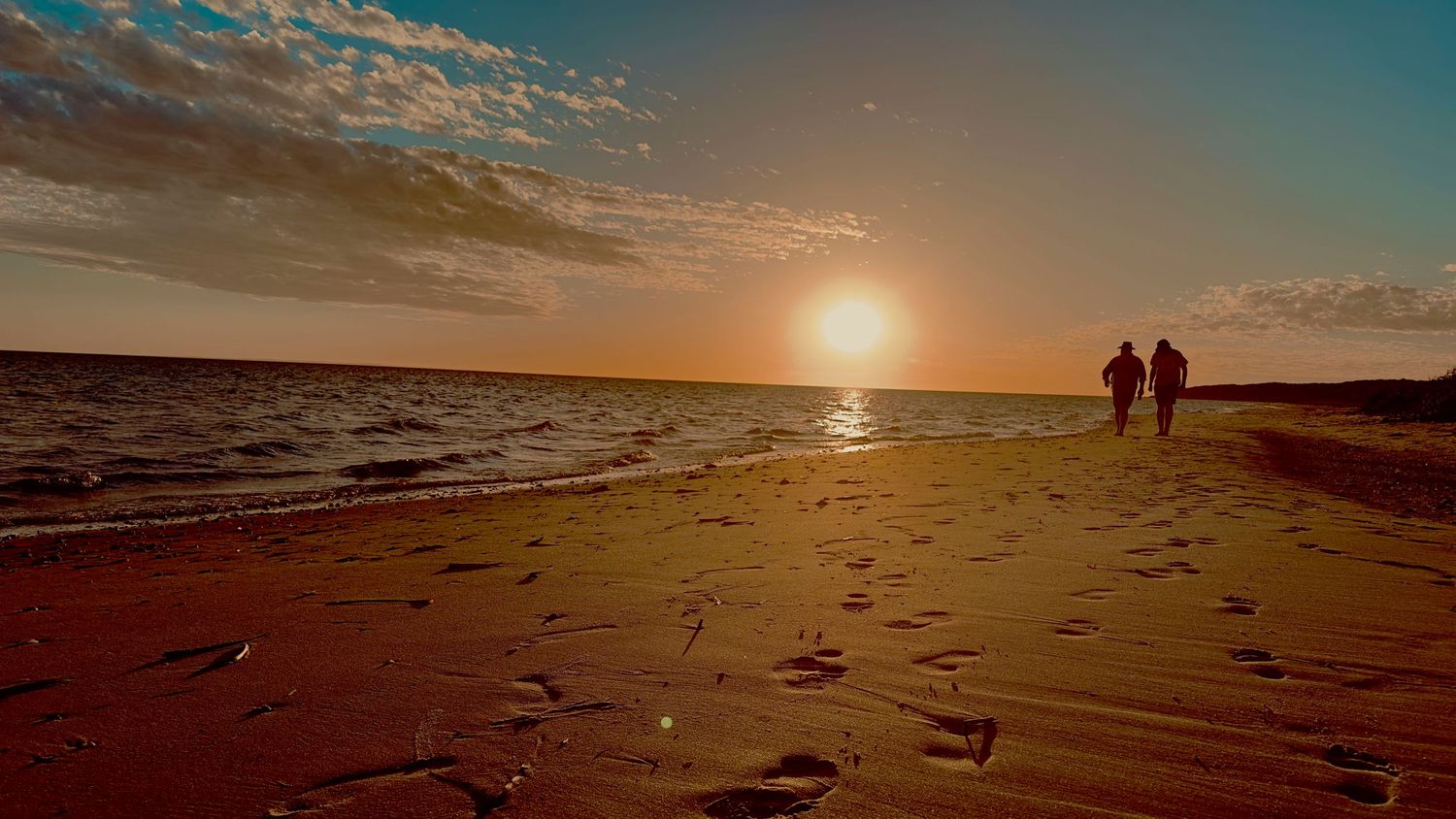 Two silhouettes walk along a sandy beach at golden sunset with footprints in the sand and calm ocean waves.
