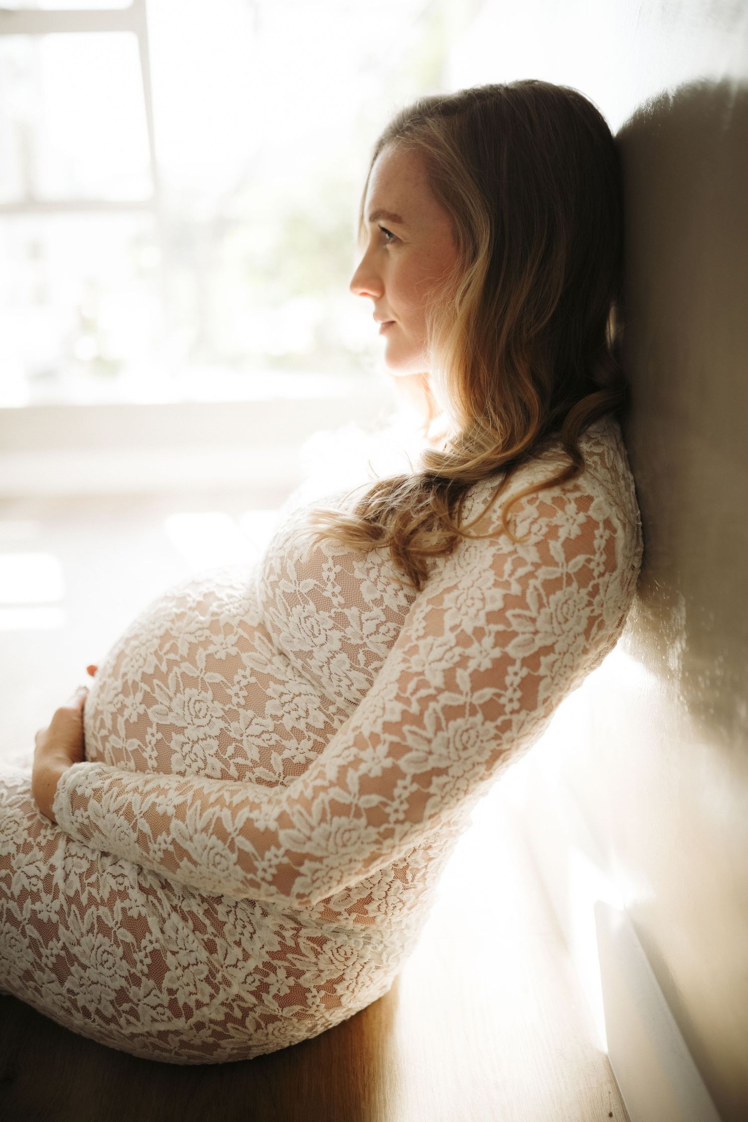 Pregnant woman in white lace dress seated by window, Sydney maternity photography