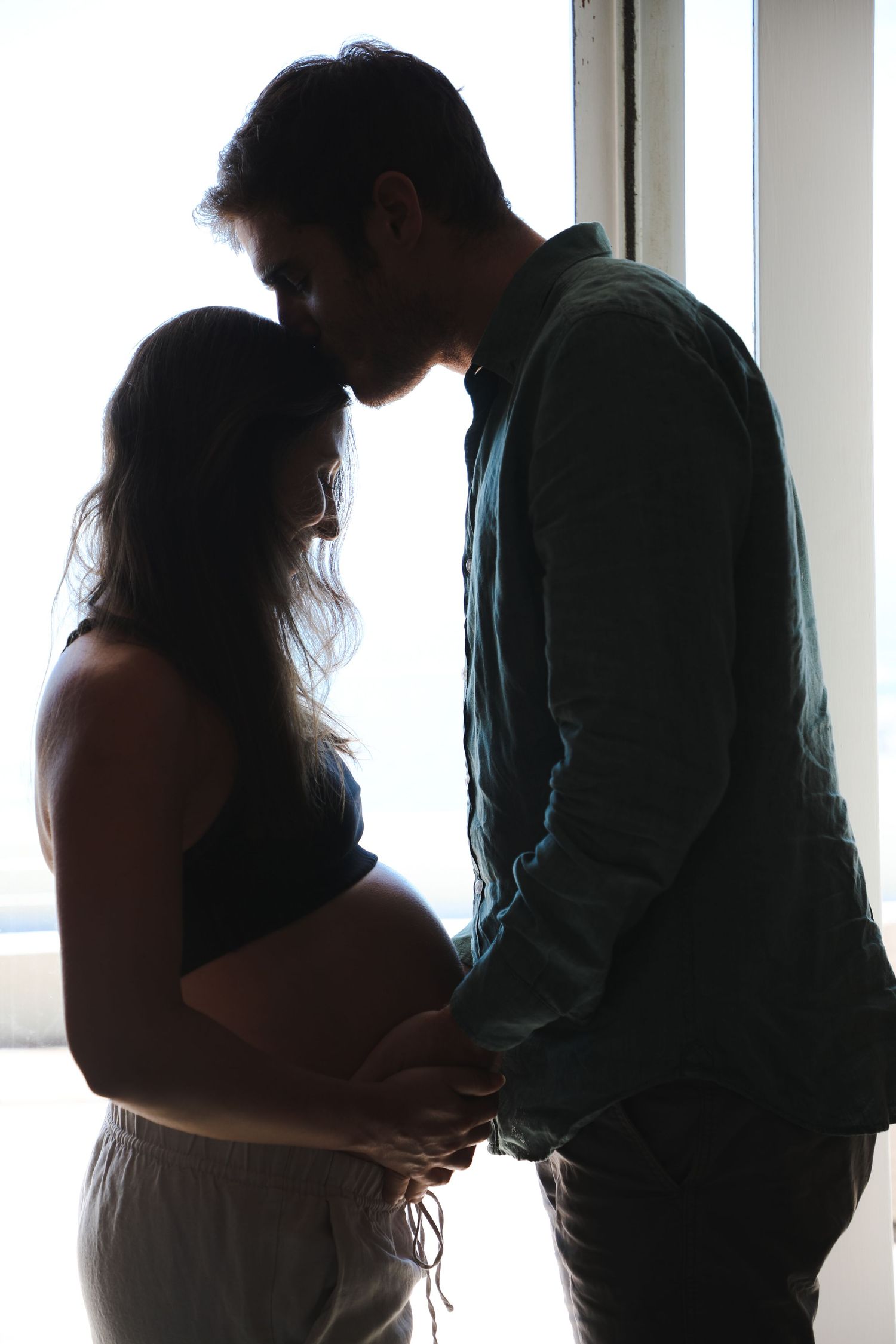 Couple sharing intimate moment forehead to forehead and holding her belly in window light, Sydney