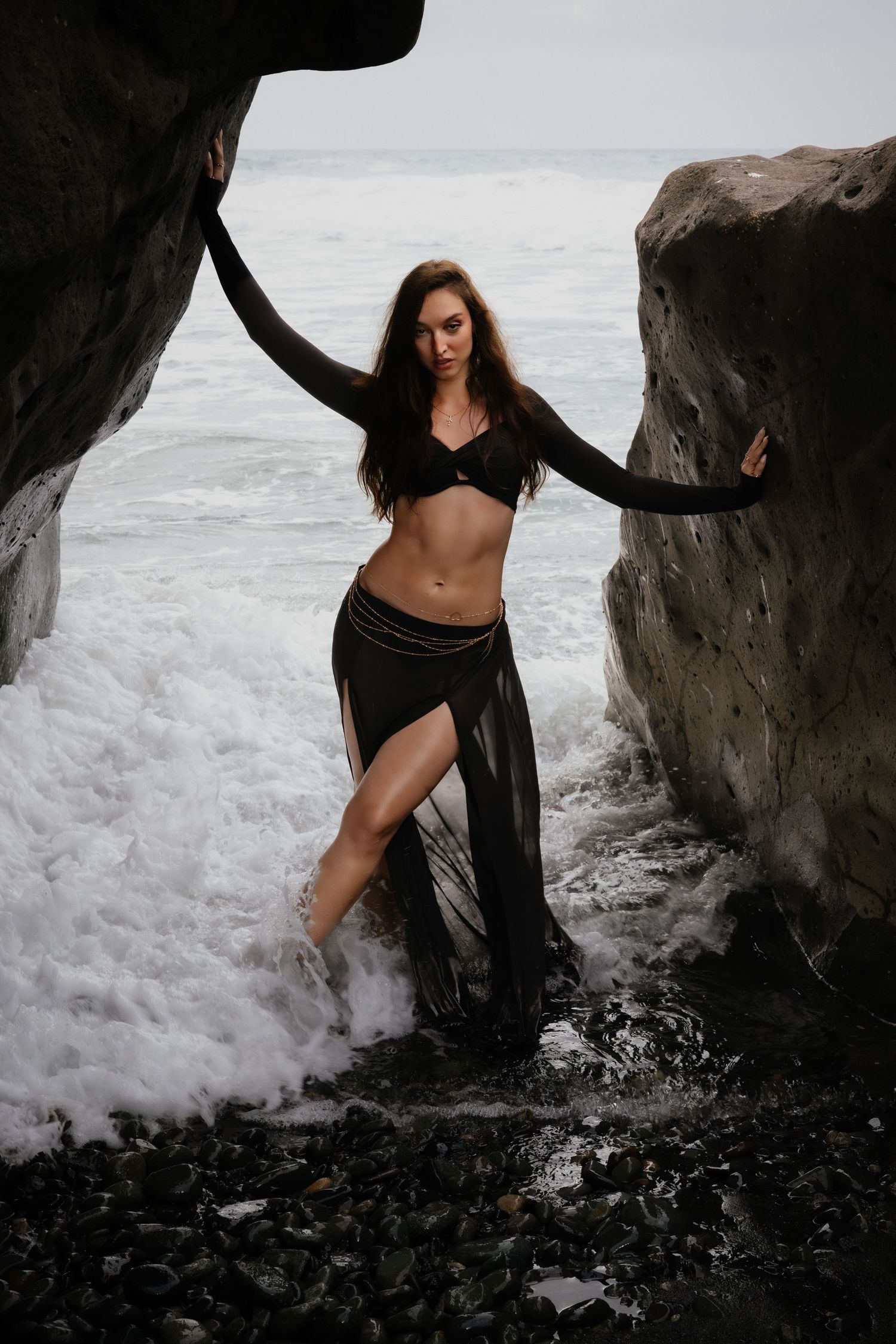 Fashion portrait of a model in black fashion wear standing between rocks in crashing sea waves, by Anya MacLaren Photography