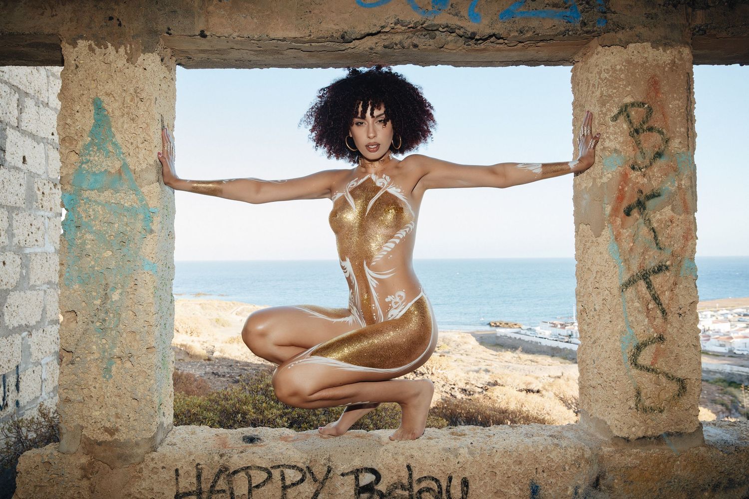 Gold glitter body paint portrait of a model crouching in a ruined stone window overlooking the coast, by Anya MacLaren Photography