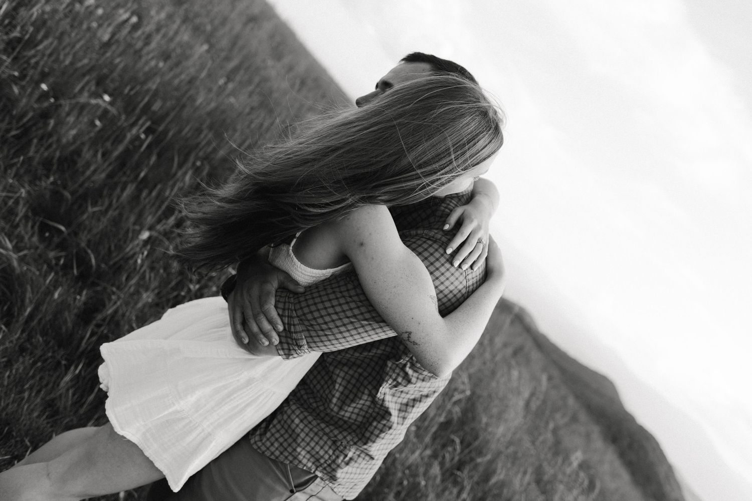 Black and white photo of a couple embracing tenderly in a field, woman's long hair flowing as they hug.