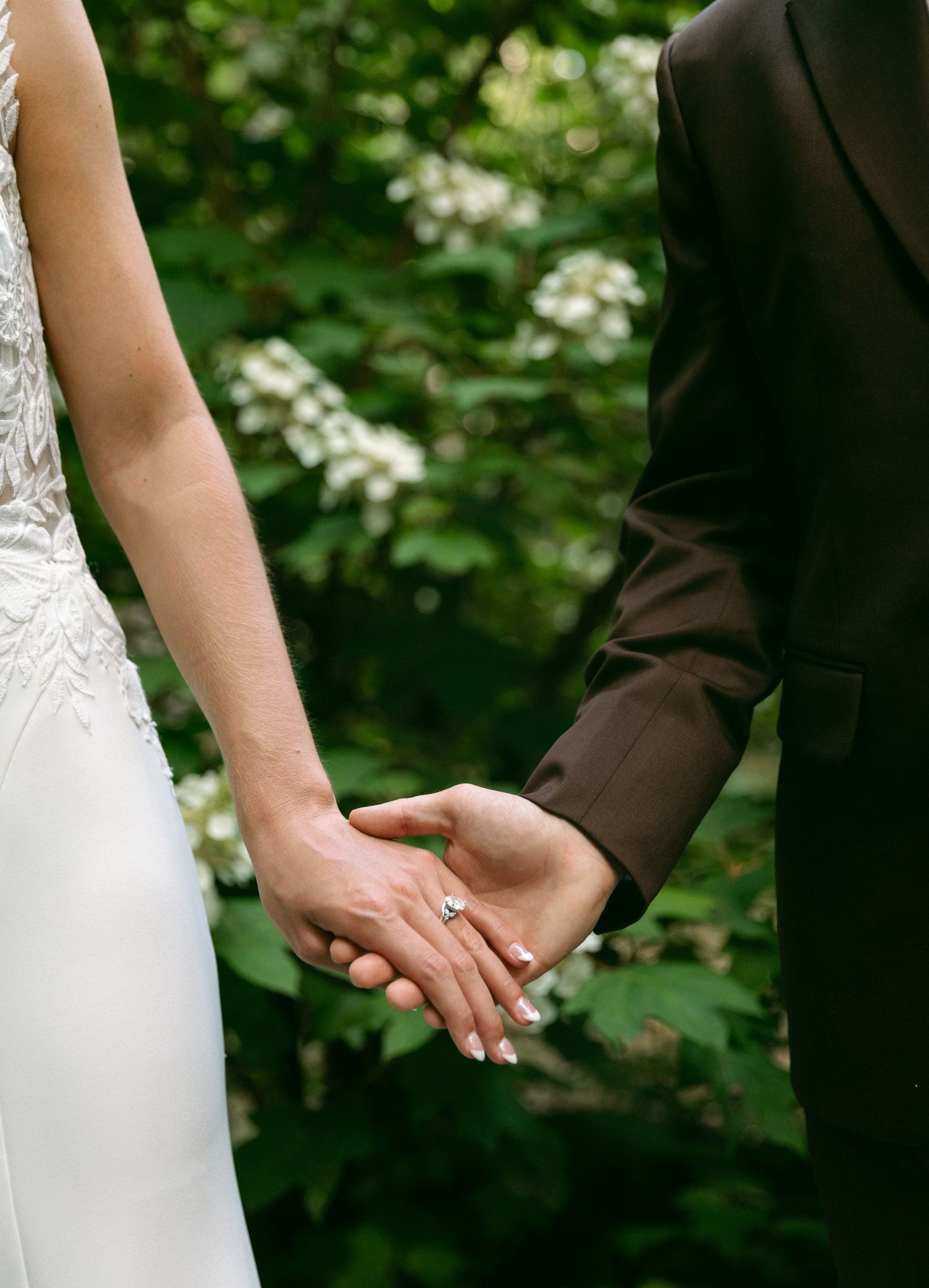 Bride and groom holding hands outdoors, showing engagement ring, wearing wedding dress and dark suit with green foliage background.