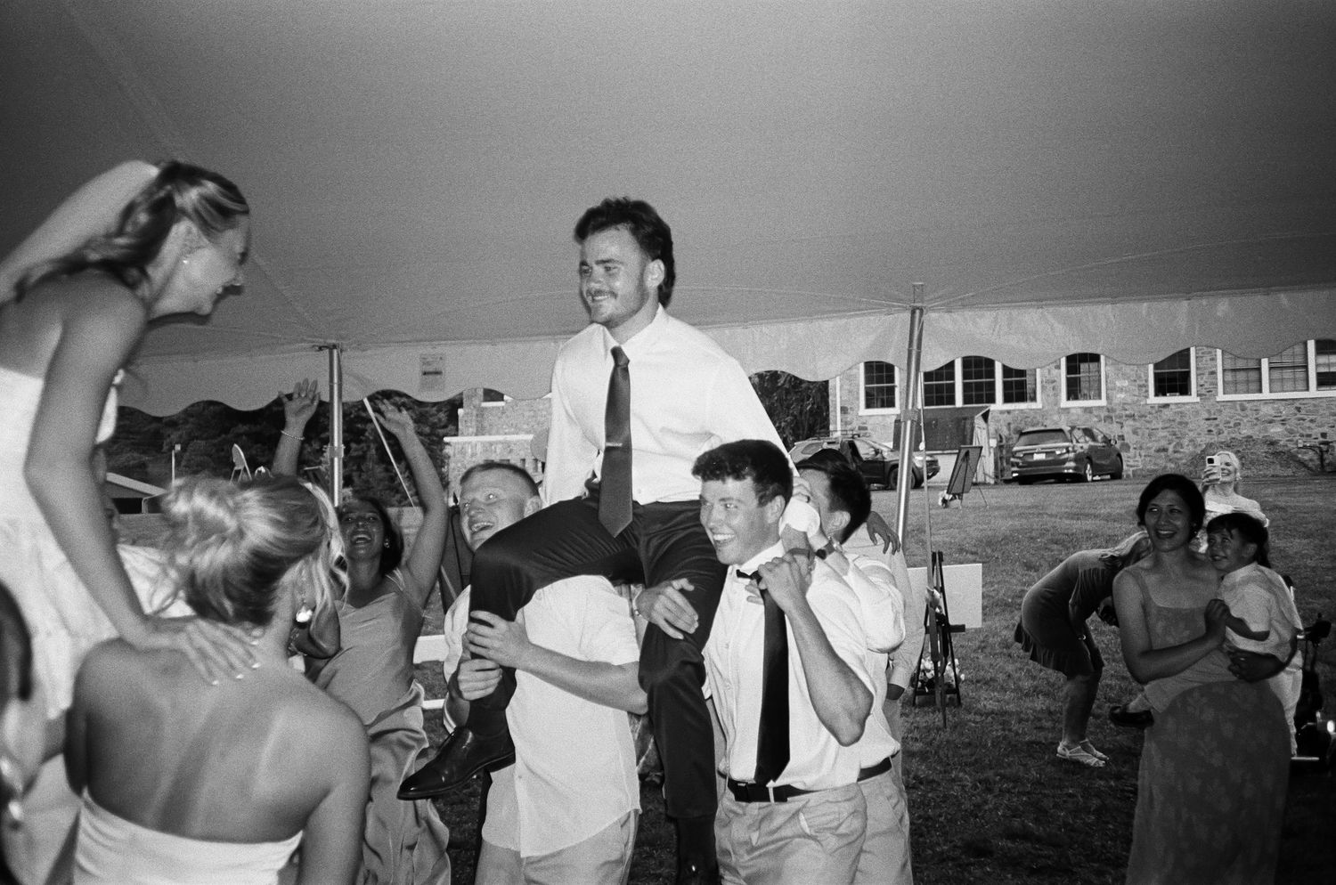 Black and white wedding photo of groom being lifted by groomsmen outdoors under a tent.
