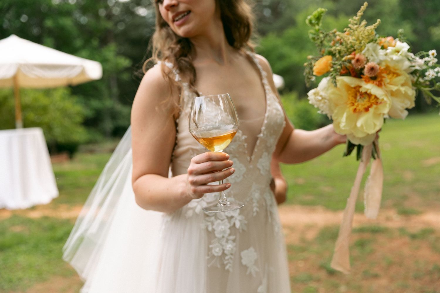 Bride in lace wedding gown holding a wine glass and yellow floral bouquet outdoors at a garden wedding.