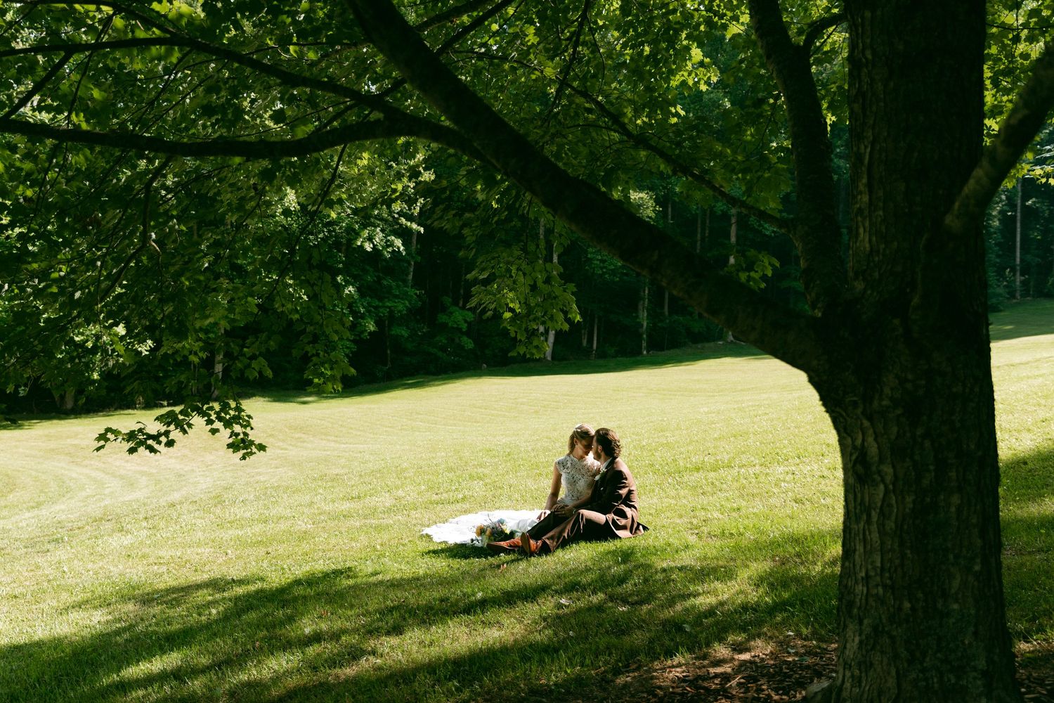 Bride and groom sitting together under large shady tree on lush green lawn at outdoor wedding.