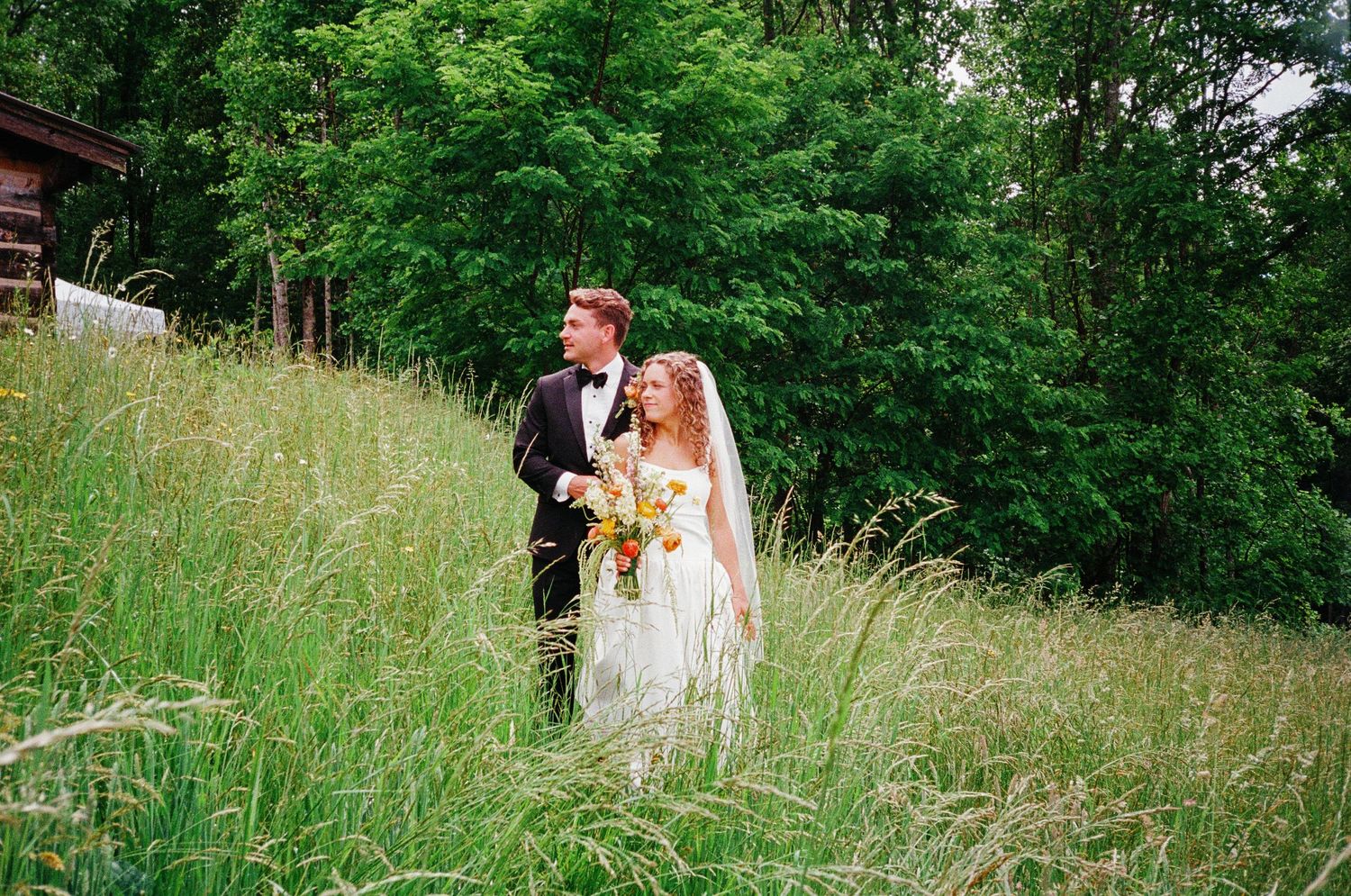 Bride and groom standing in tall green meadow grass near rustic barn, bride holding colorful bouquet on wedding day.