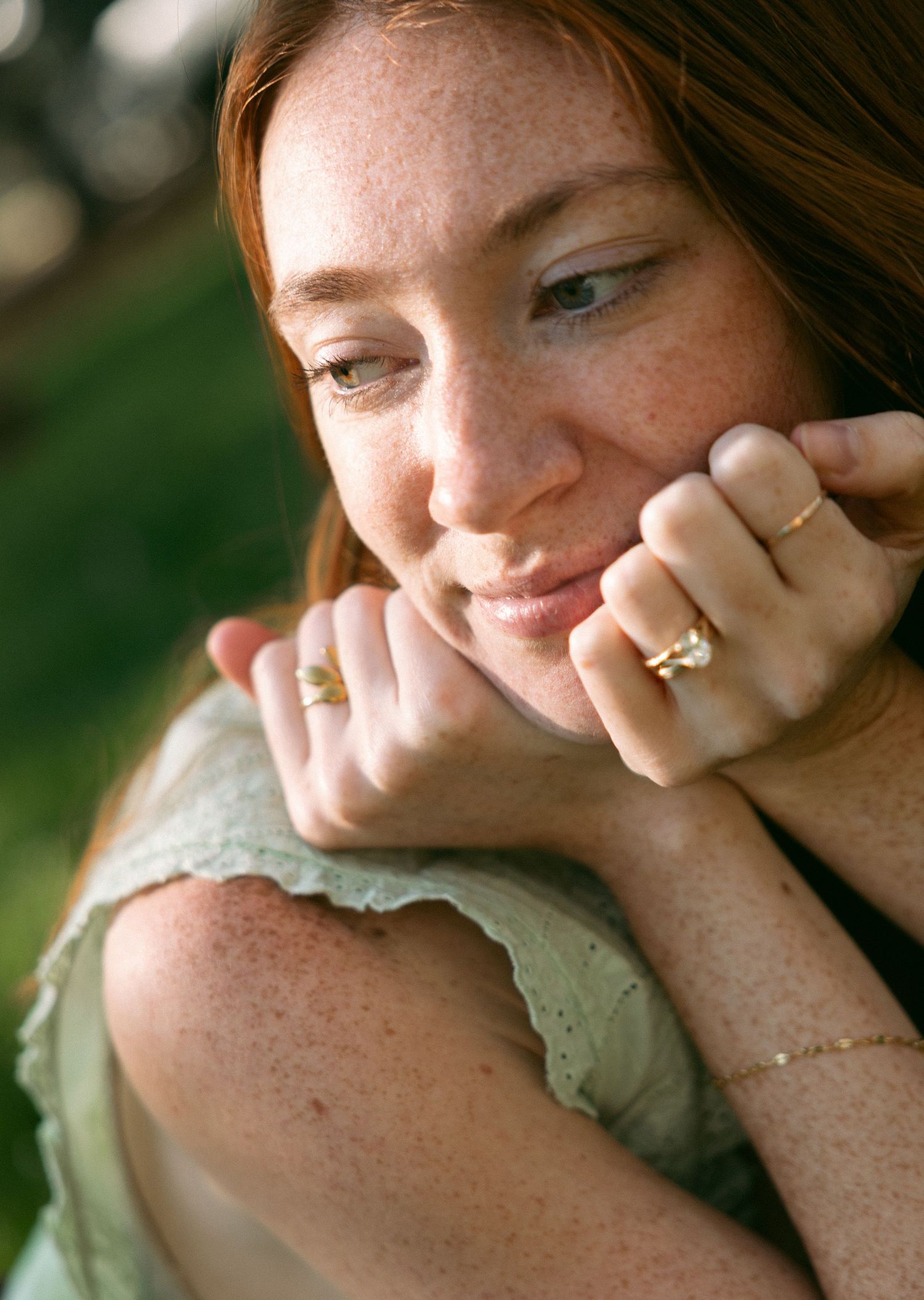 A freckled redhead woman rests her chin on her hands, wearing gold rings, with a soft dreamy expression outdoors.