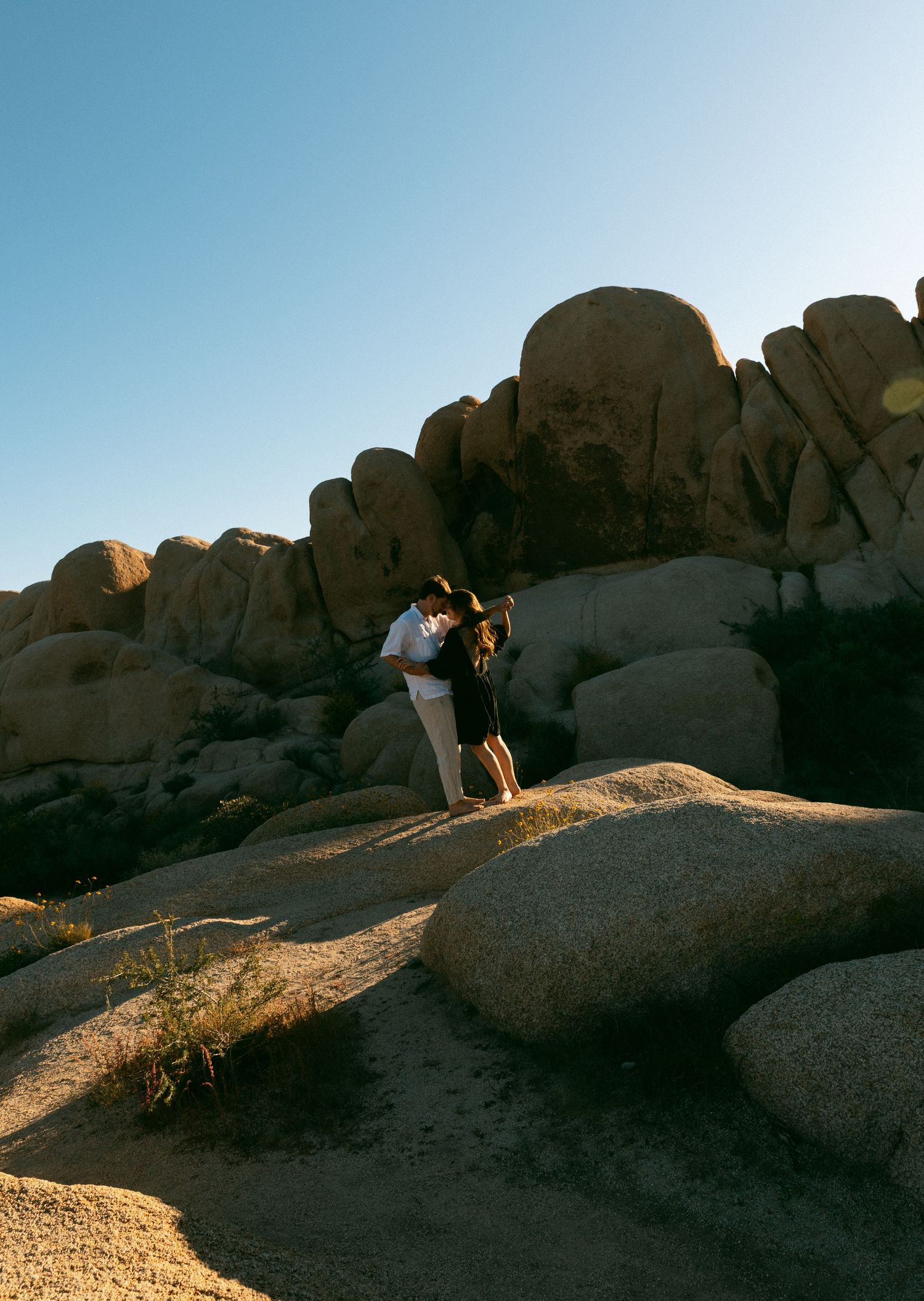 Couple embracing romantically on rocky boulders at Joshua Tree National Park under a clear blue sky.