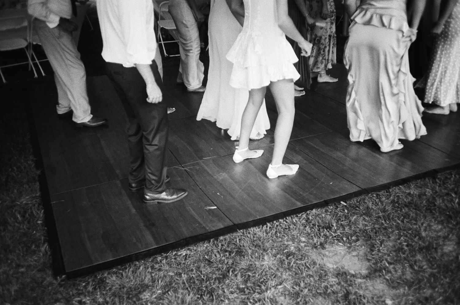 Black and white photo of people dancing barefoot on a wooden dance floor at an outdoor wedding reception.