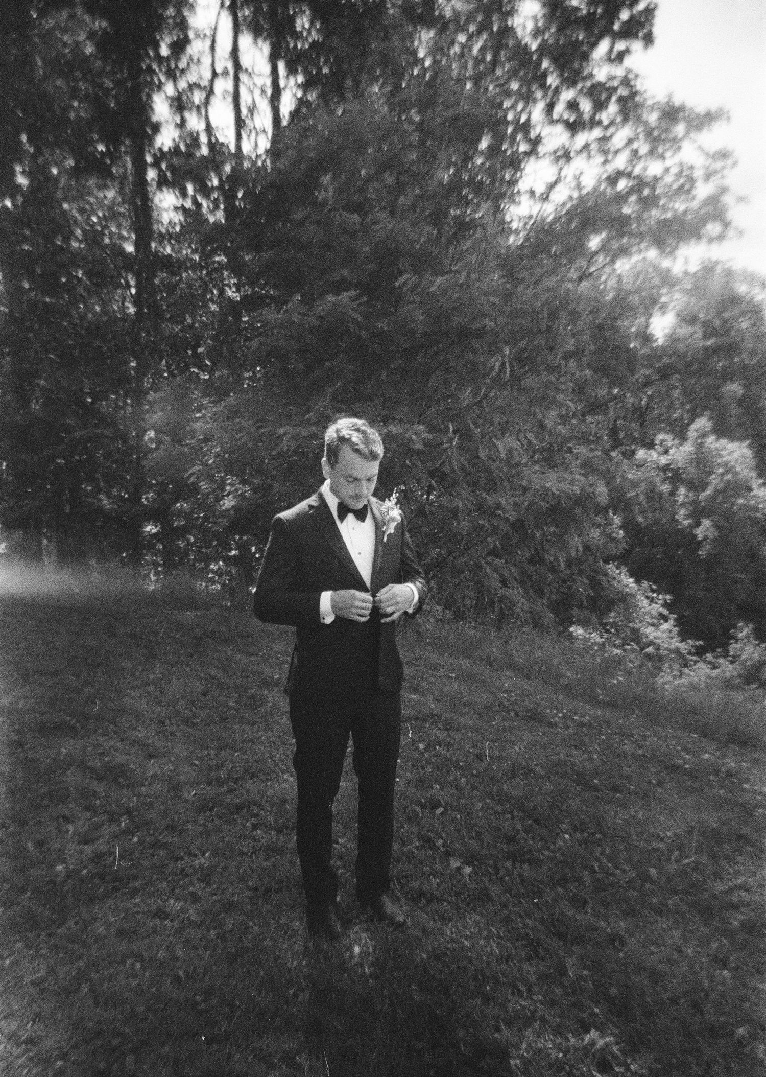 A groom in a black tuxedo and bow tie buttons his jacket while standing on a grassy lawn surrounded by trees.