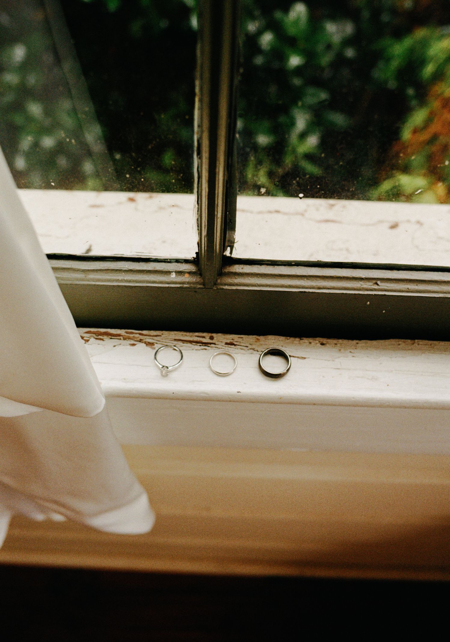 Three wedding rings resting on a weathered white window sill with a lace veil beside them.