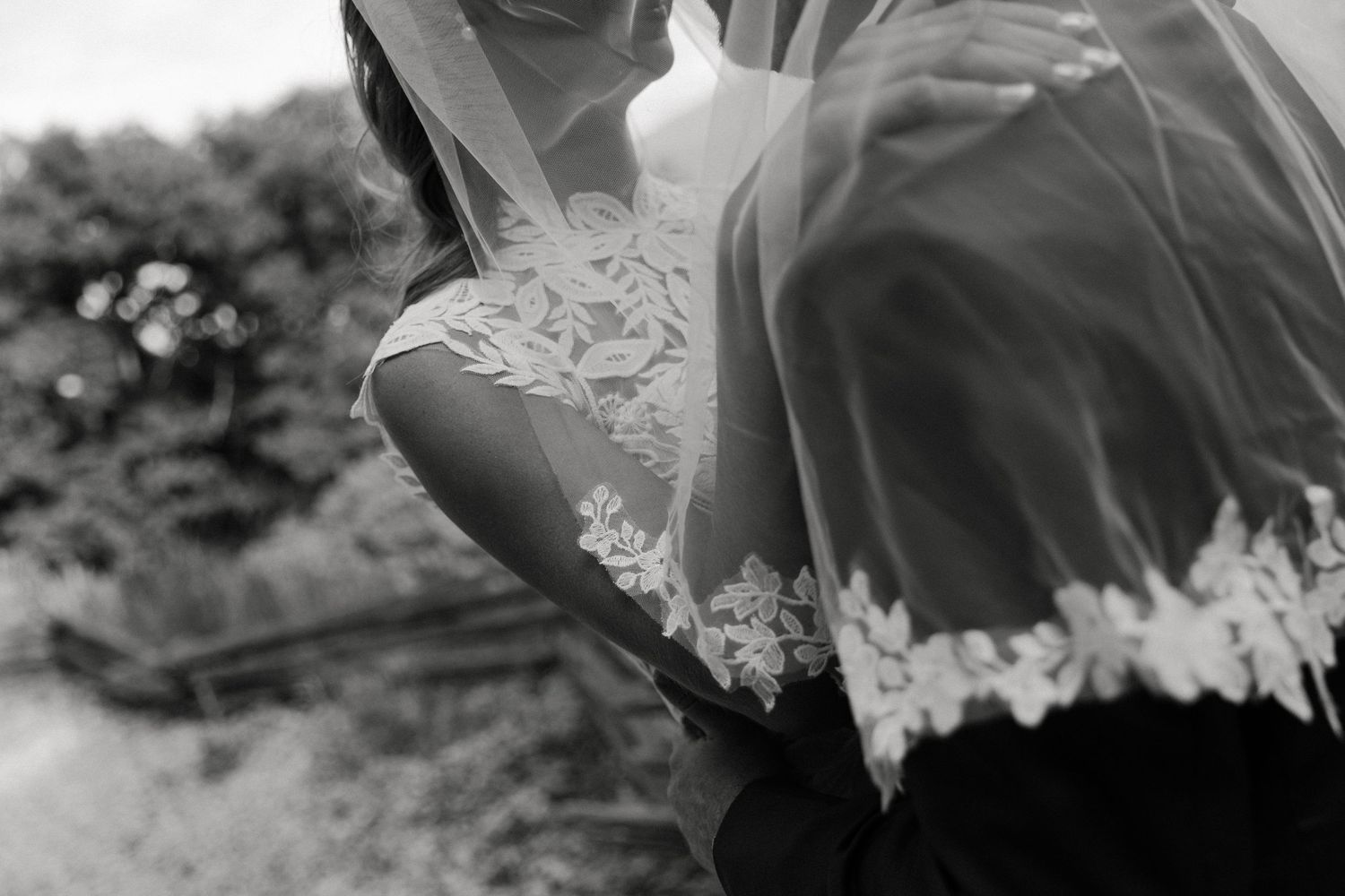 Black and white close-up of bride in lace-trimmed veil embracing groom at romantic outdoor wedding.